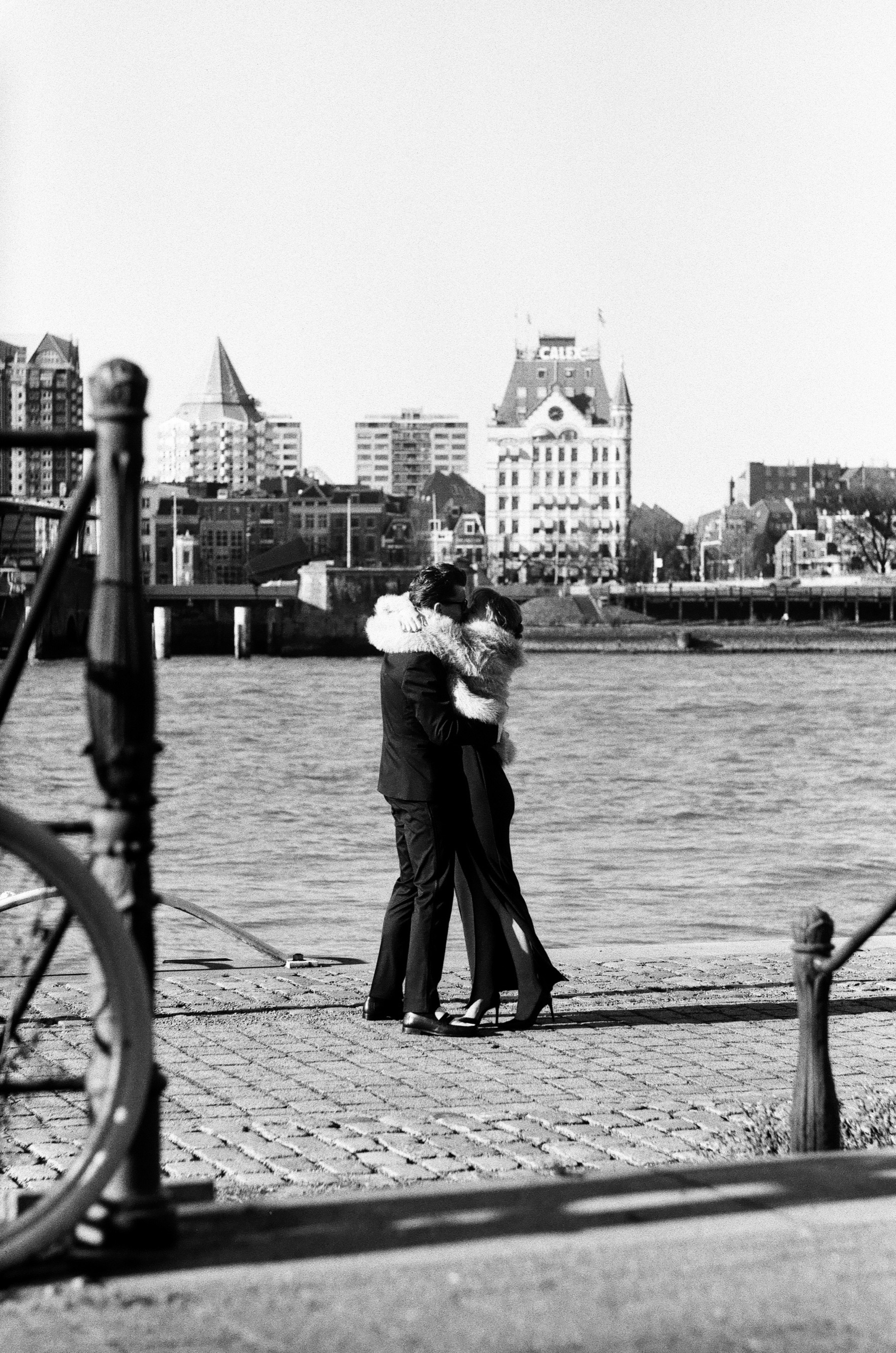 Couple hugging on a cobbled quay of the Maas with Het Witte Huis across the water, a proposal photo session in Rotterdam on black and white film