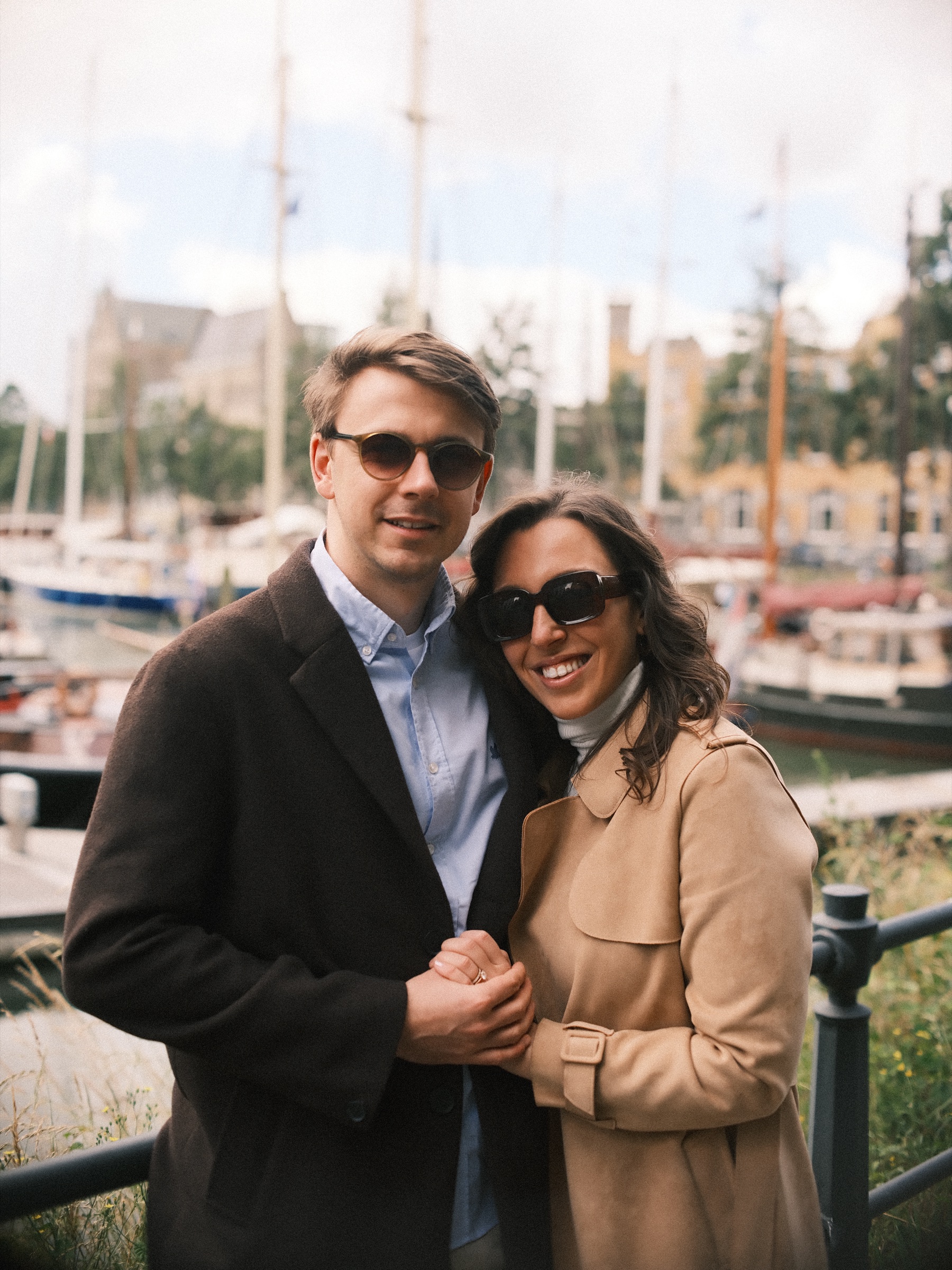 Smiling couple in sunglasses hugging by the Veerhaven railing, sailing ship masts behind them, their engagement shoot in the Scheepvaartkwartier