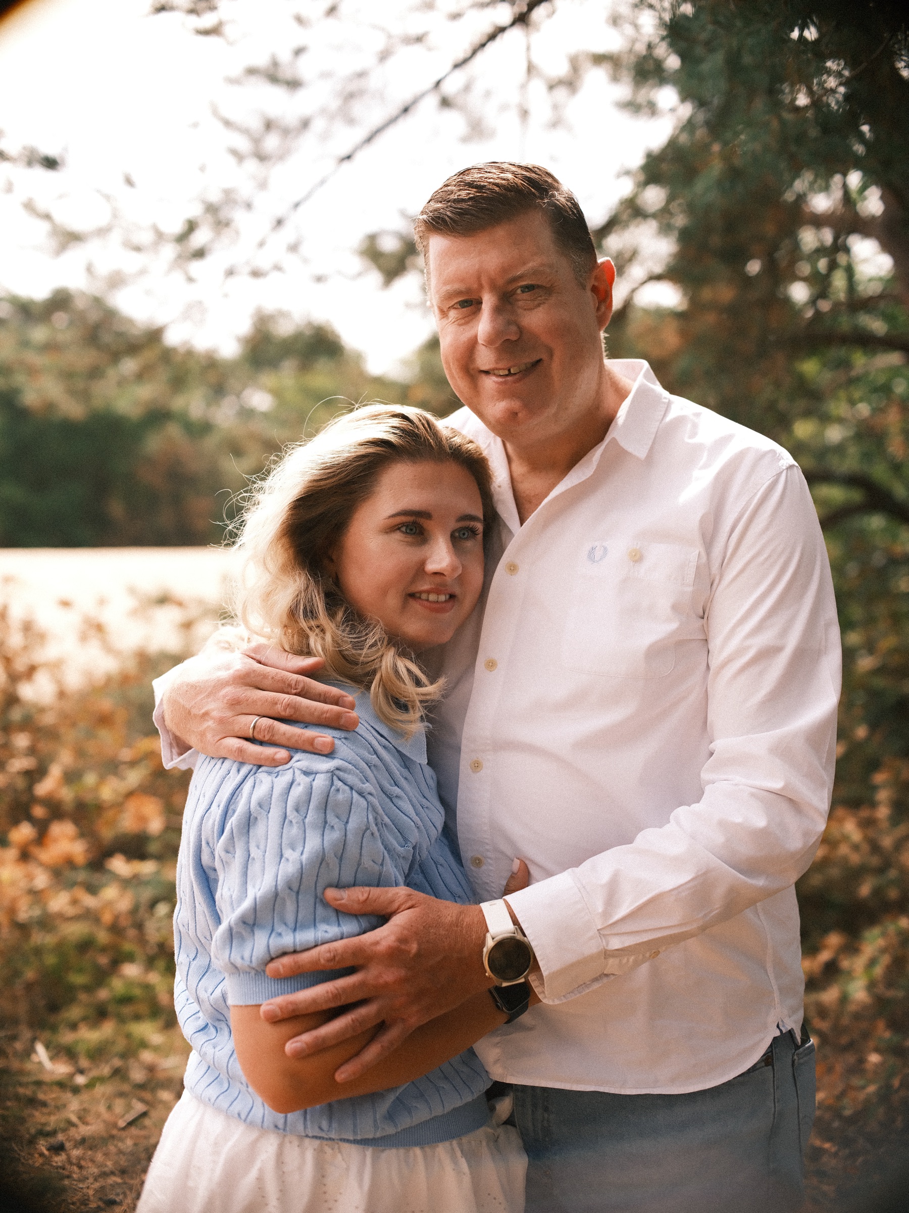 Man in a white shirt holding his partner in a blue knit top close, her head against his chest, orange-brown shrubs behind