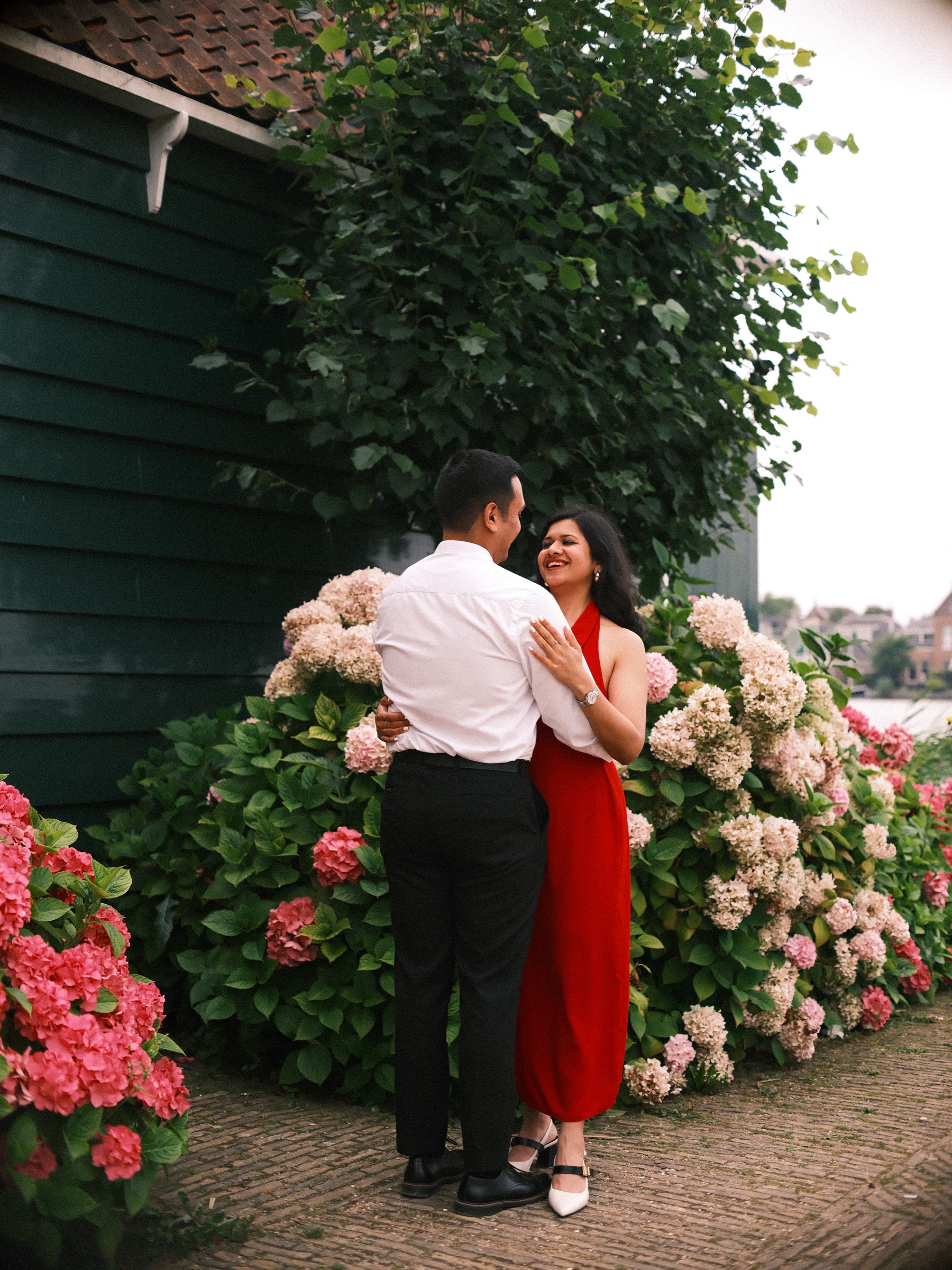 Couple embracing among pink and cream hydrangeas beside a dark green wooden house, the river Zaan and the far bank just visible behind