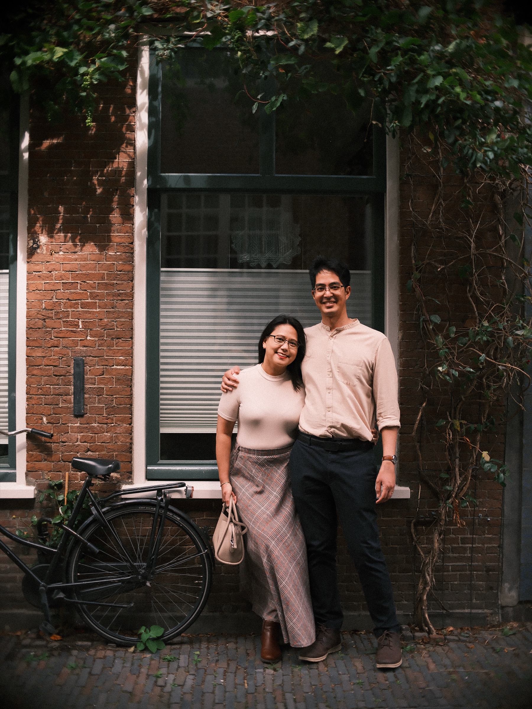 Couple arm in arm under ivy by a window with a lowered blind, an old black bicycle leaning against the brick wall beside them