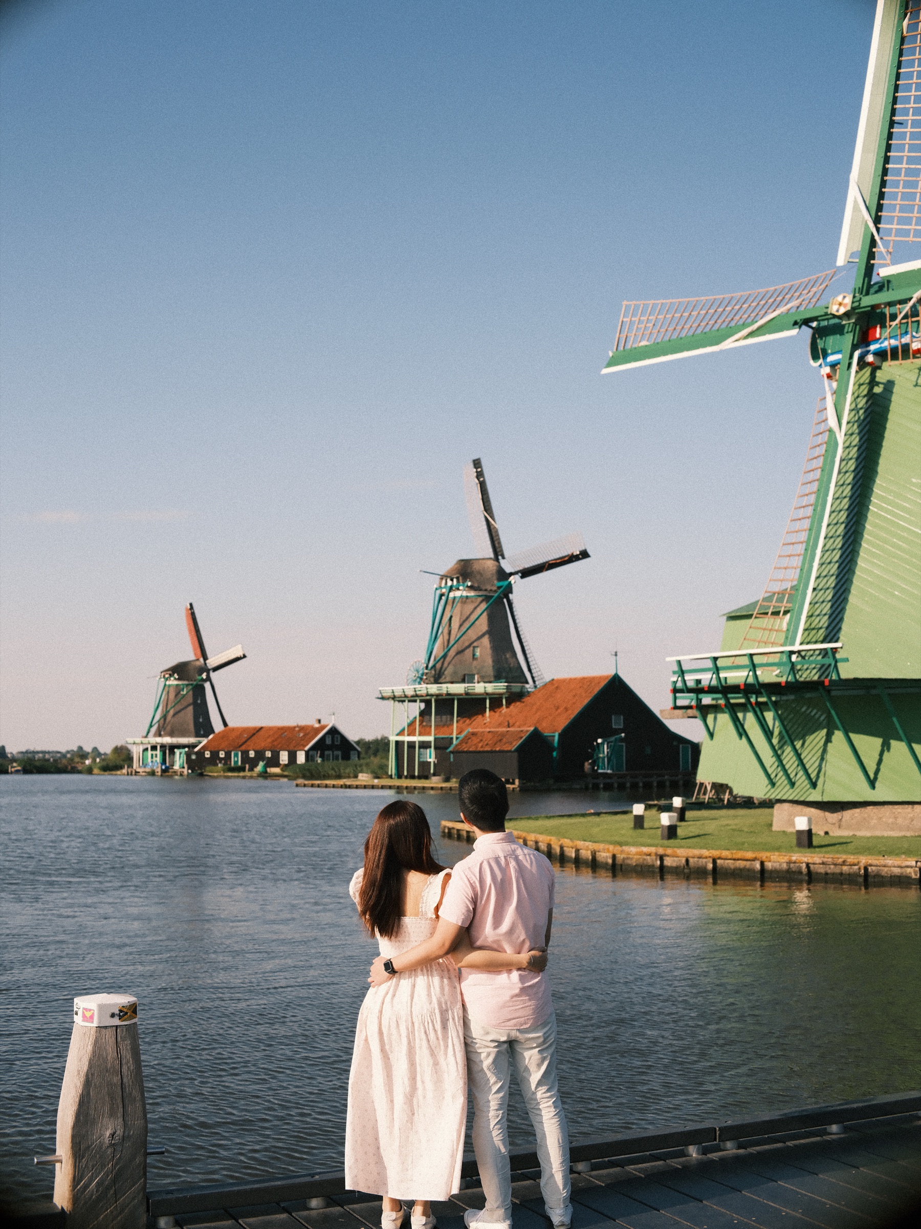 Back view of an engaged couple holding each other on a wooden jetty, looking across the Zaan at a row of windmills