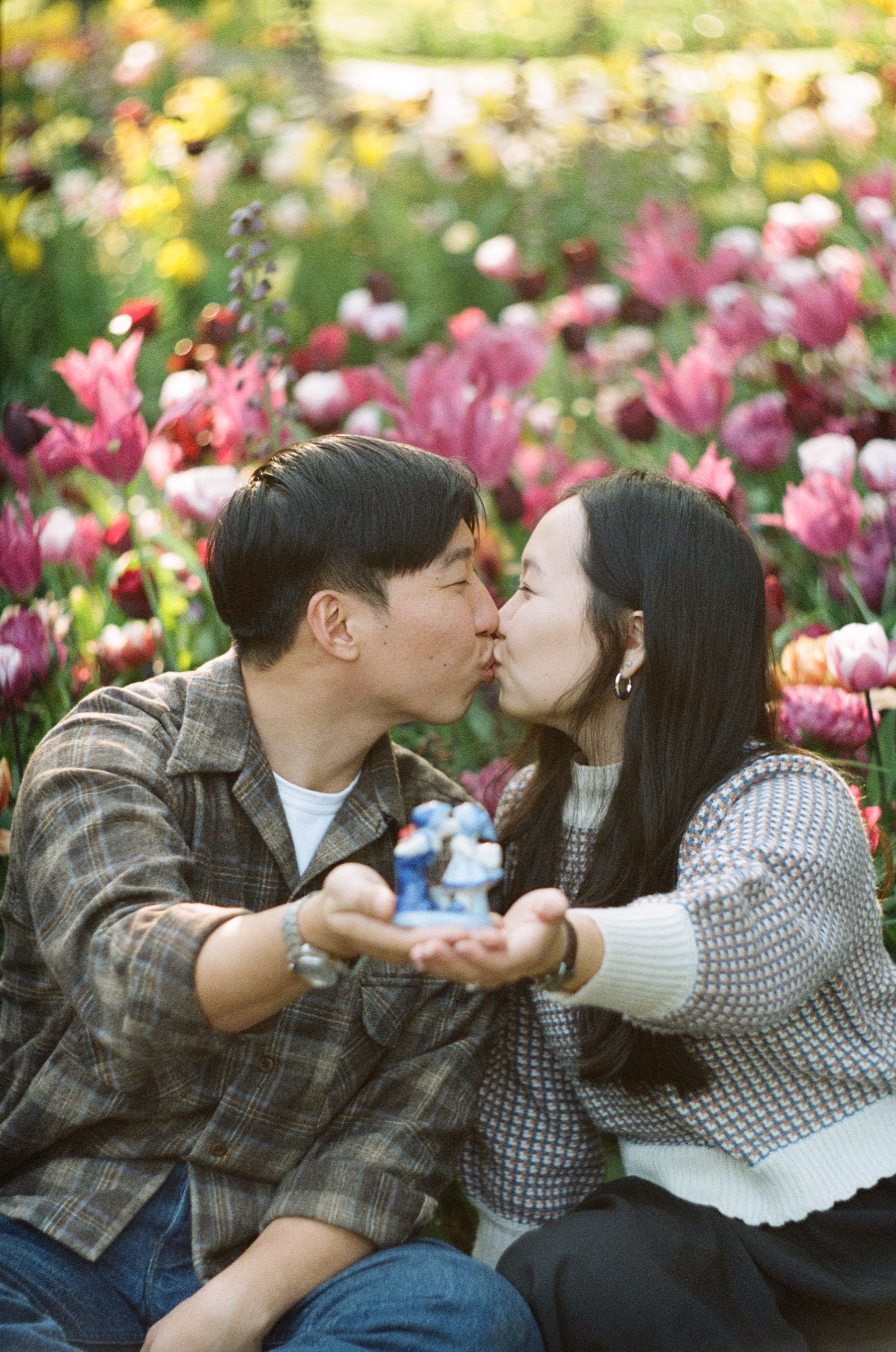 Couple kissing while they hold the little blue-and-white figurine of a kissing couple between them among pink tulips