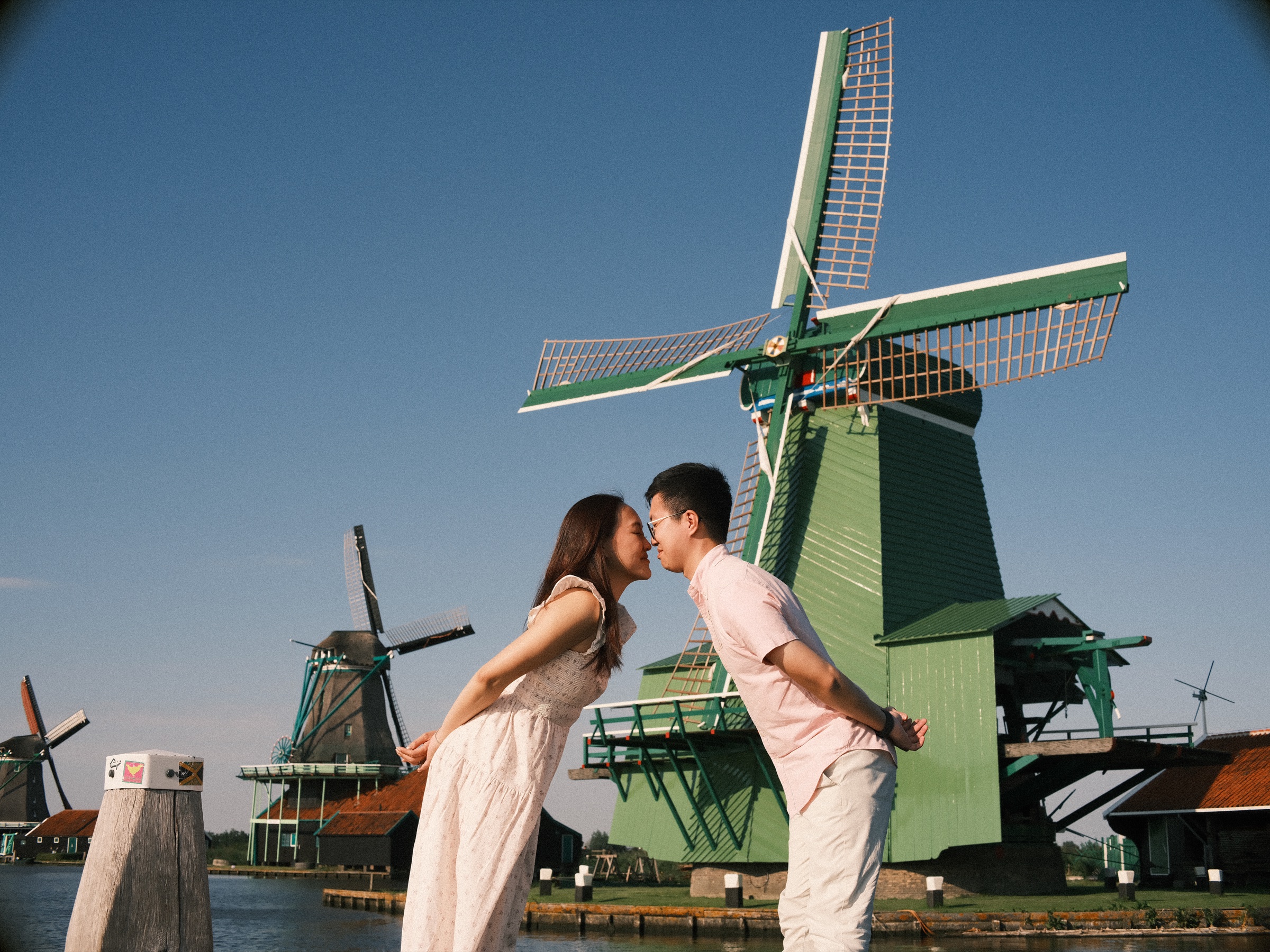 Engaged couple leaning in for a kiss with their hands behind their backs, a green windmill and the Zaan behind them at the Zaanse Schans