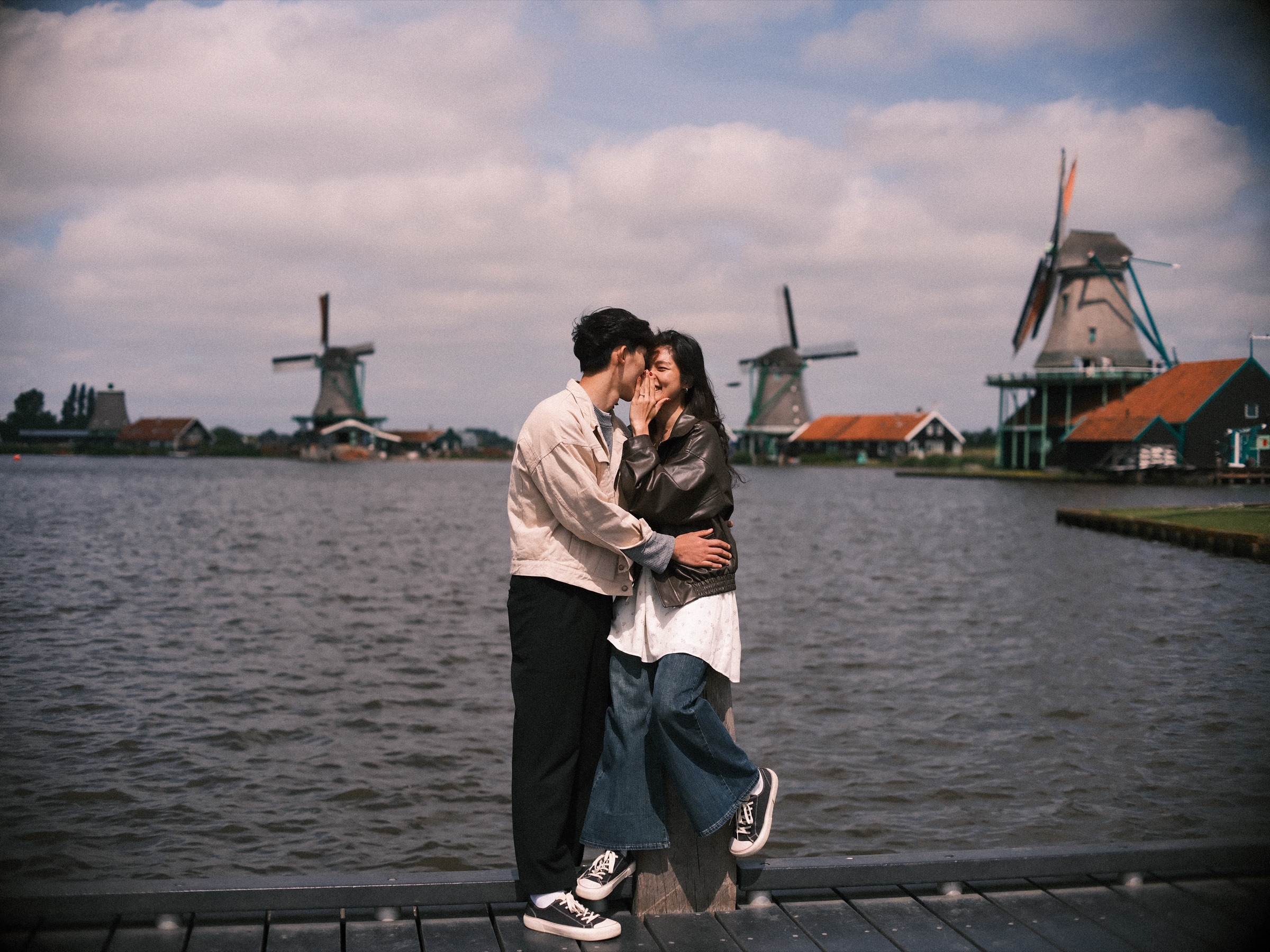 Man kissing the cheek of his laughing fiancée on a wooden jetty over the Zaan, three windmills of the Zaanse Schans across the water