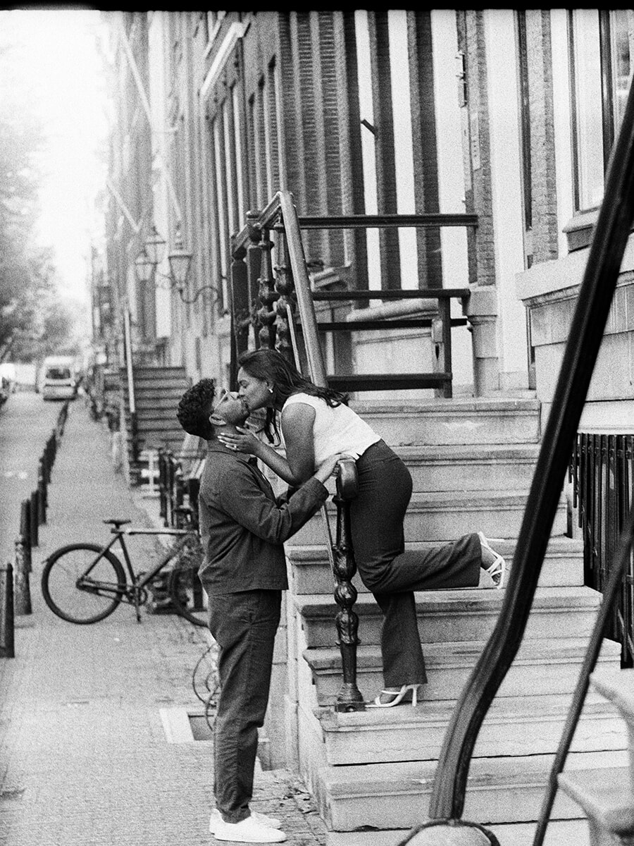 On stone steps in Amsterdam, a woman leans down to kiss her partner, one leg bent behind her, during an analog couple photoshoot