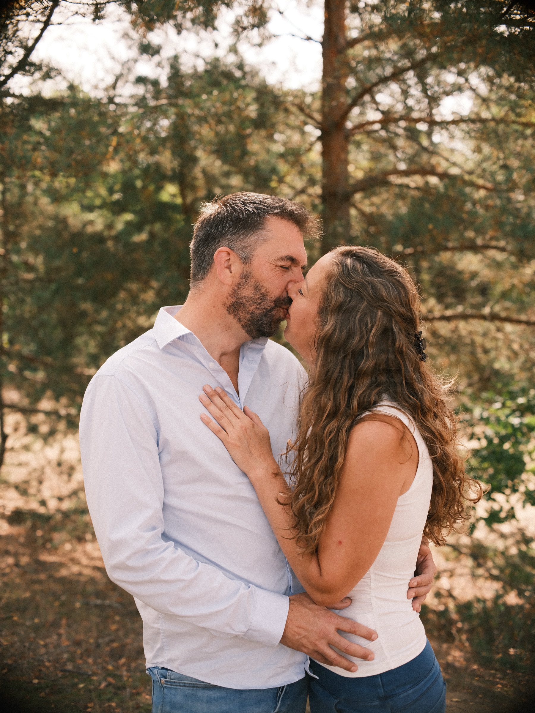 Bearded man in a white shirt kissing his partner with long curly hair under the pines, her hand resting on his chest