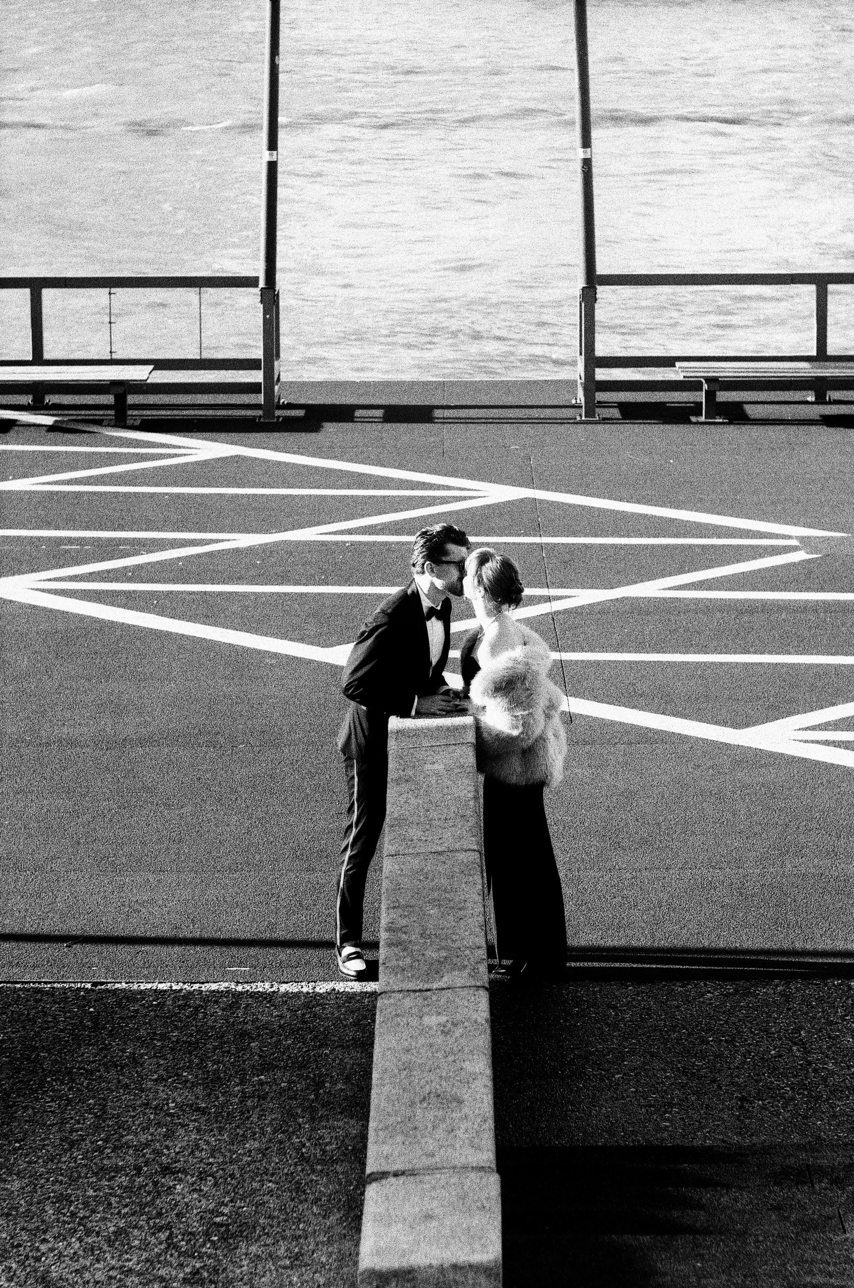 Seen from above, the couple kisses in the middle of white painted lines on an empty quay, the river glittering behind them