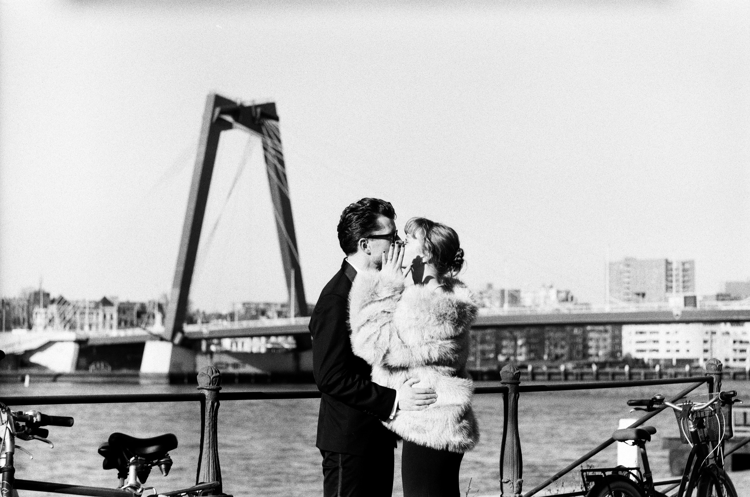 Kiss by the railing of the Maas in Rotterdam, her hand at her cheek, the pylon of the Willemsbrug rising behind them and bikes in front