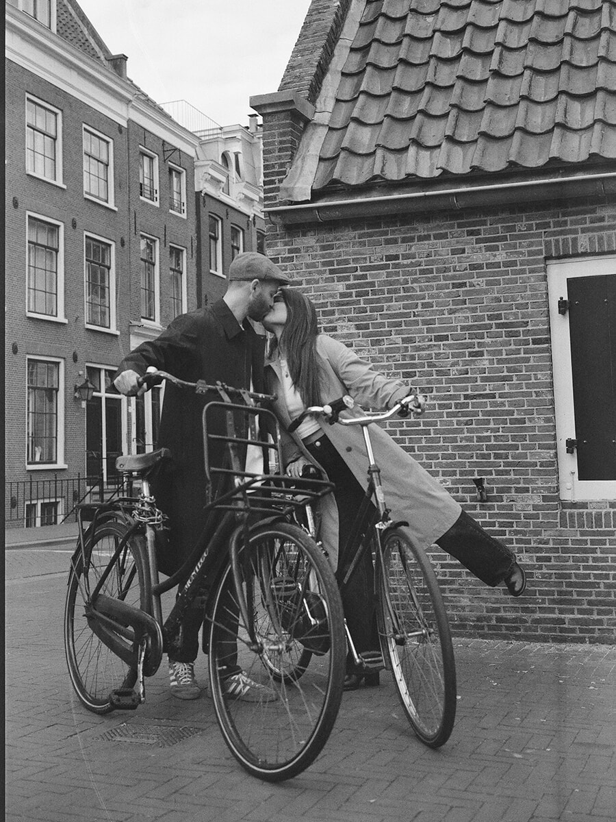 A couple kiss beside two bicycles against a brick wall in Amsterdam, her leg lifted behind her in this film portrait