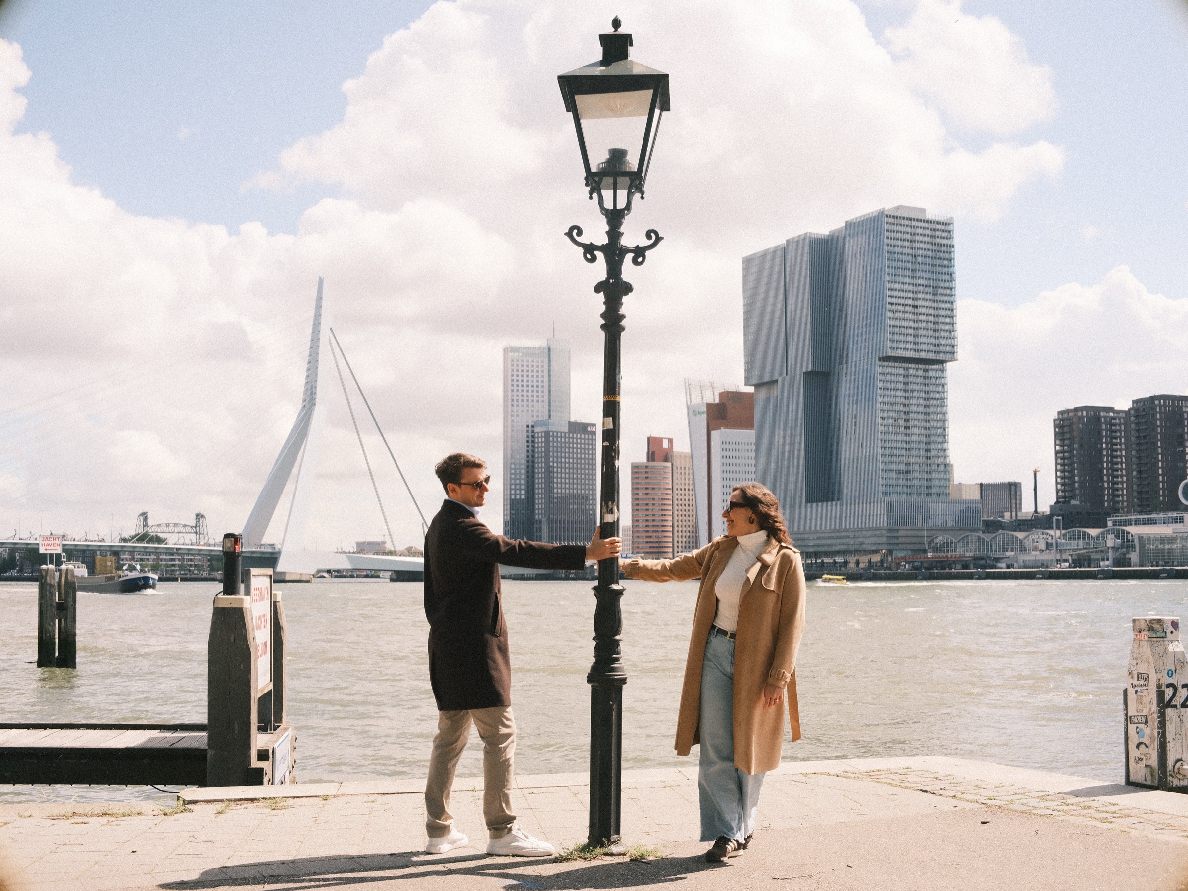 Couple in long coats holding hands around an old lamppost on the Maas quay, a photo spot in Rotterdam facing the Erasmusbrug and De Rotterdam