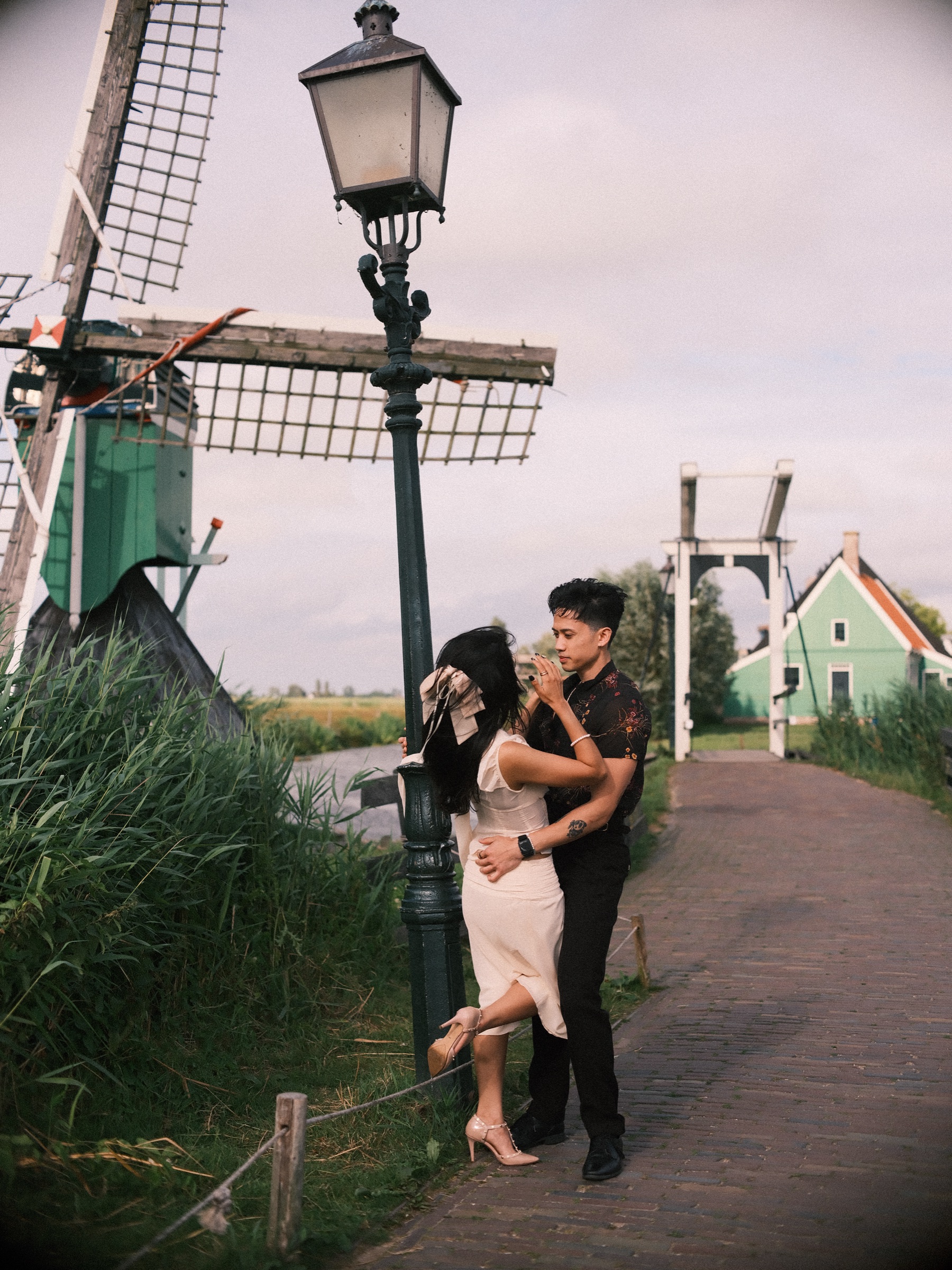 Couple holding each other by an old lamppost on a brick path, a small mill with lattice sails, tall reeds and a white drawbridge behind