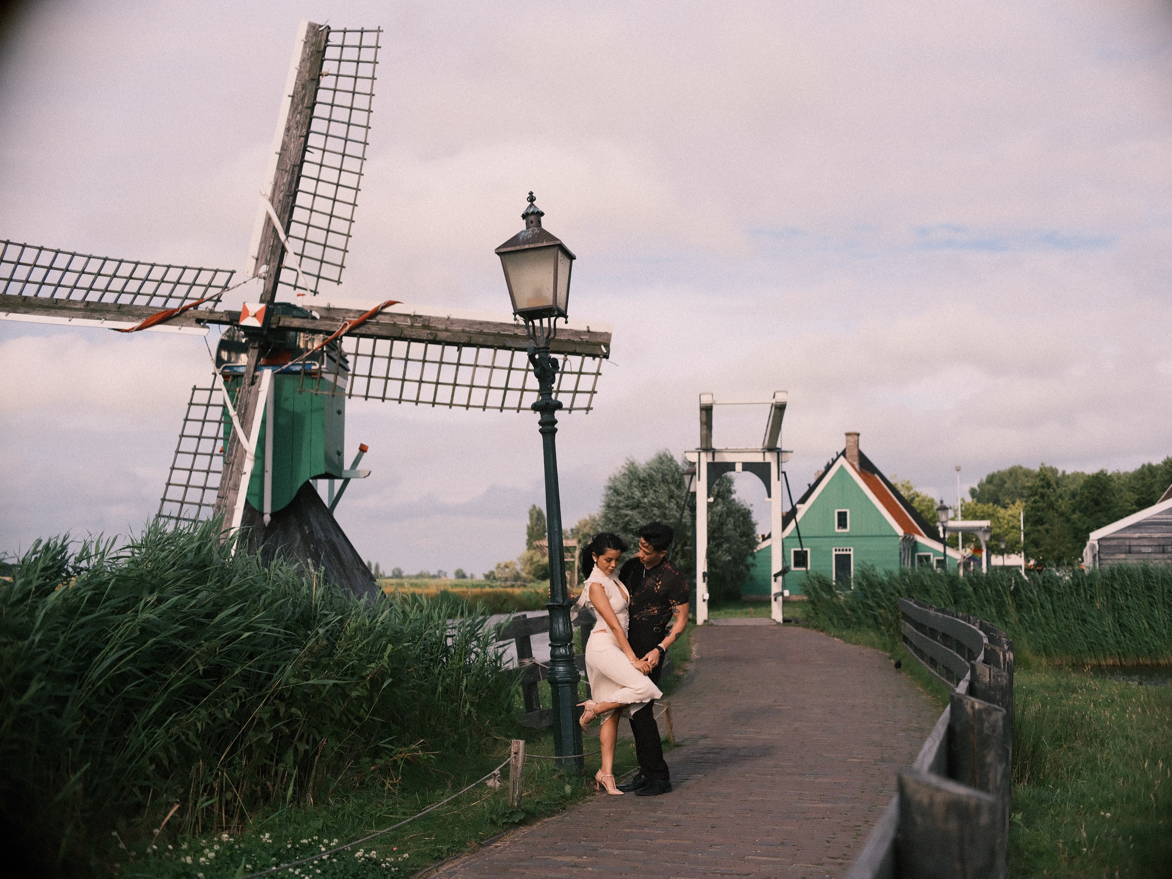 Couple standing by an old lamppost on a brick path beside a small windmill and a white drawbridge at the Zaanse Schans