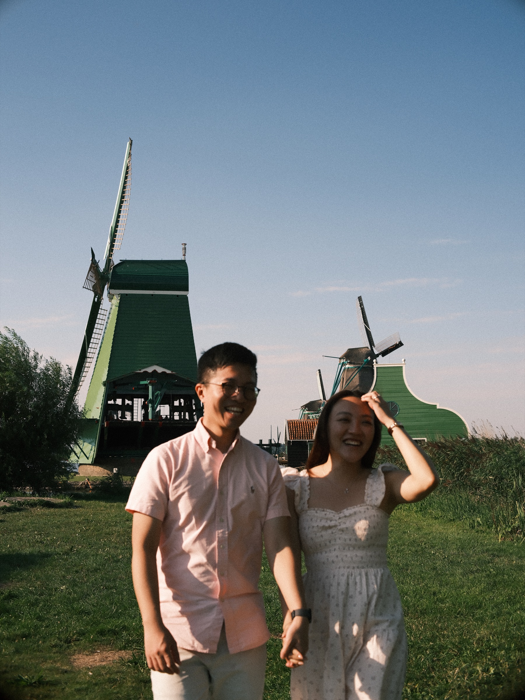 Close frame of the couple laughing in the sun, her hand holding her hair against the wind, windmills soft in the distance