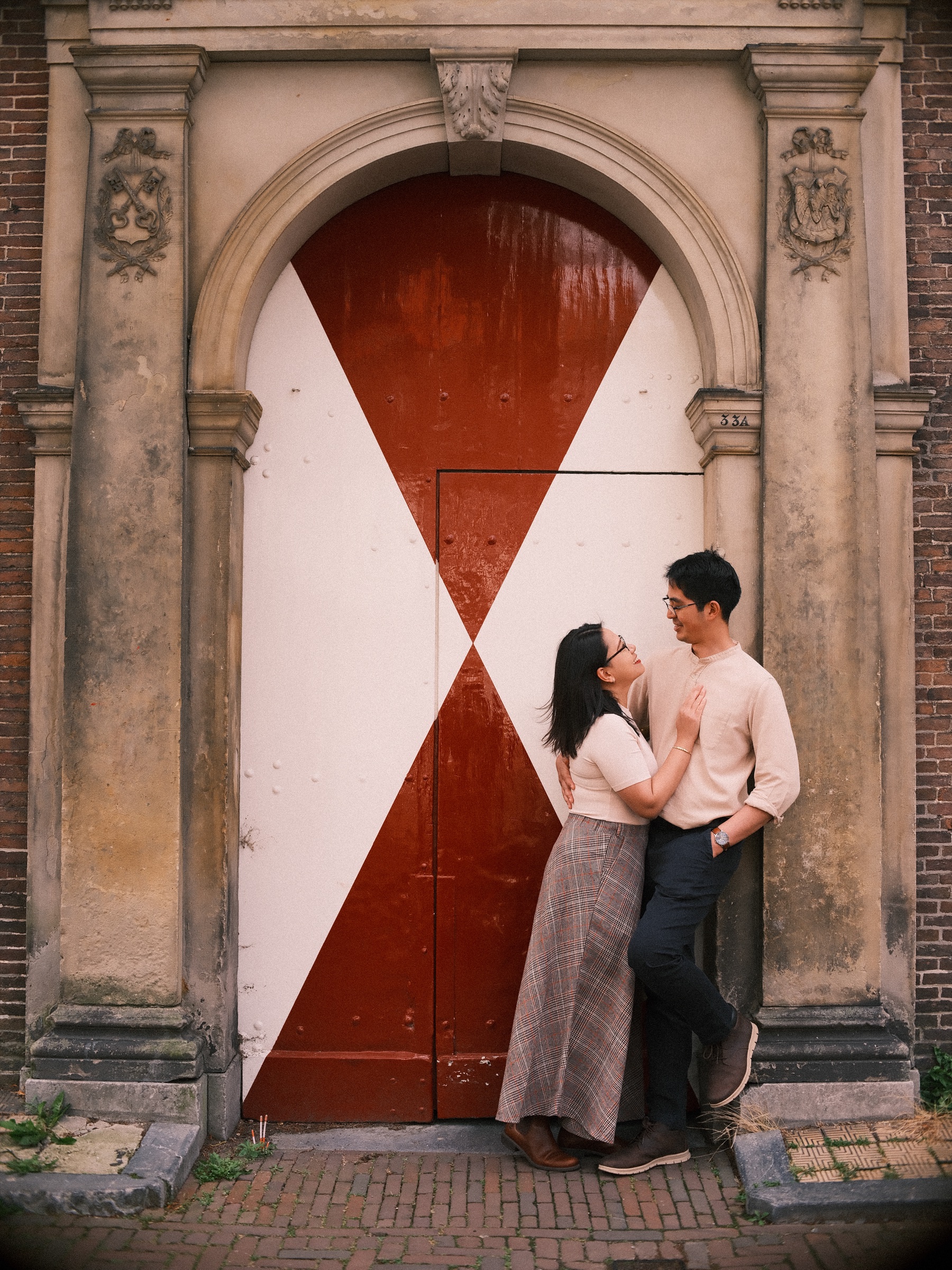 Couple laughing against an old stone gateway with red and white painted doors, his hand in his pocket and hers on his chest