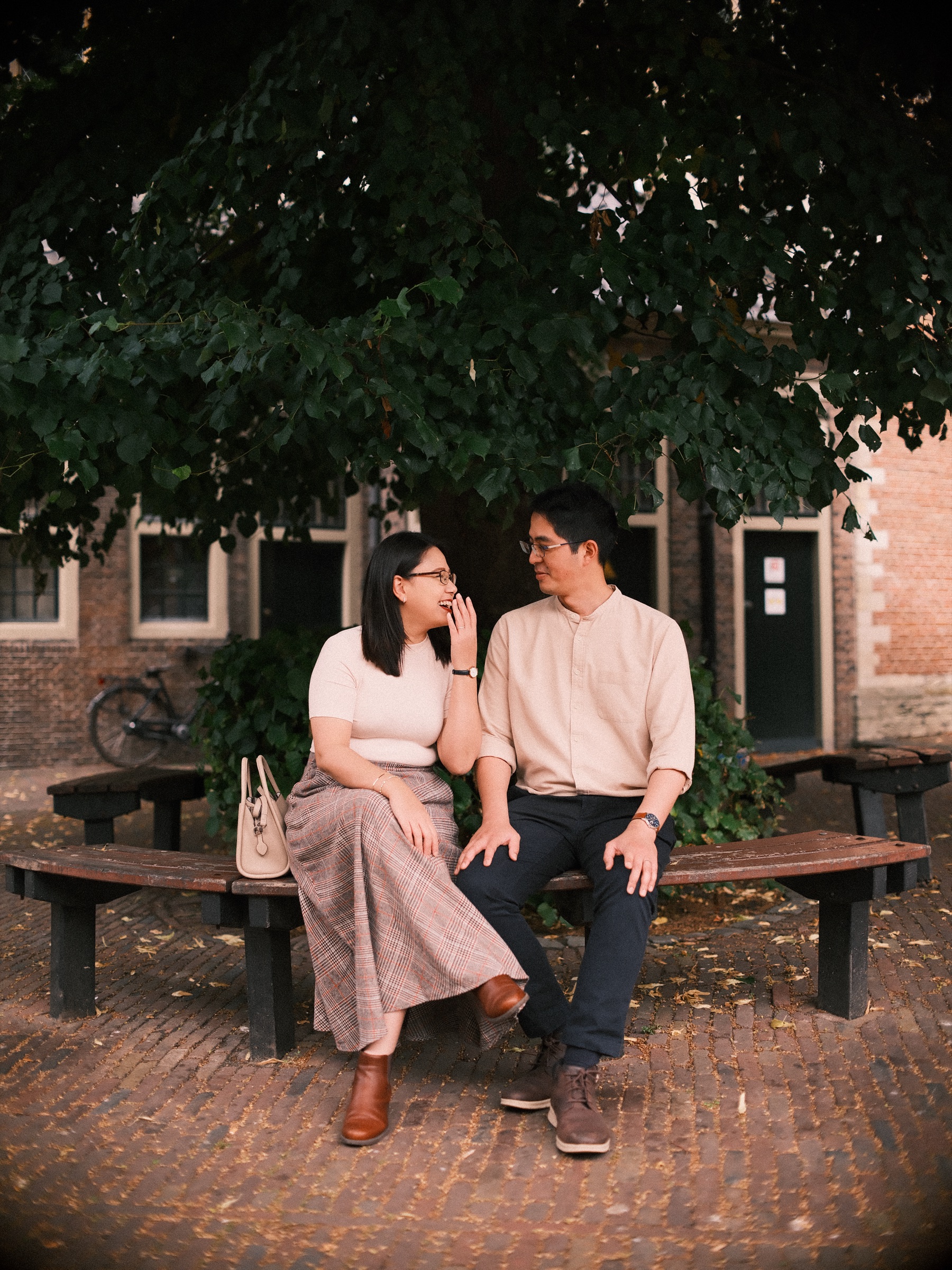 Couple on a round wooden bench in the deep shade of a tree, she covers her laugh with one hand while he grins at her