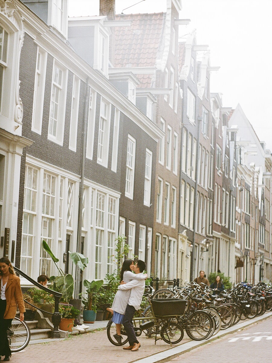 A man lifts his partner into a kiss beside parked bicycles and canal houses during an analog photoshoot in Amsterdam
