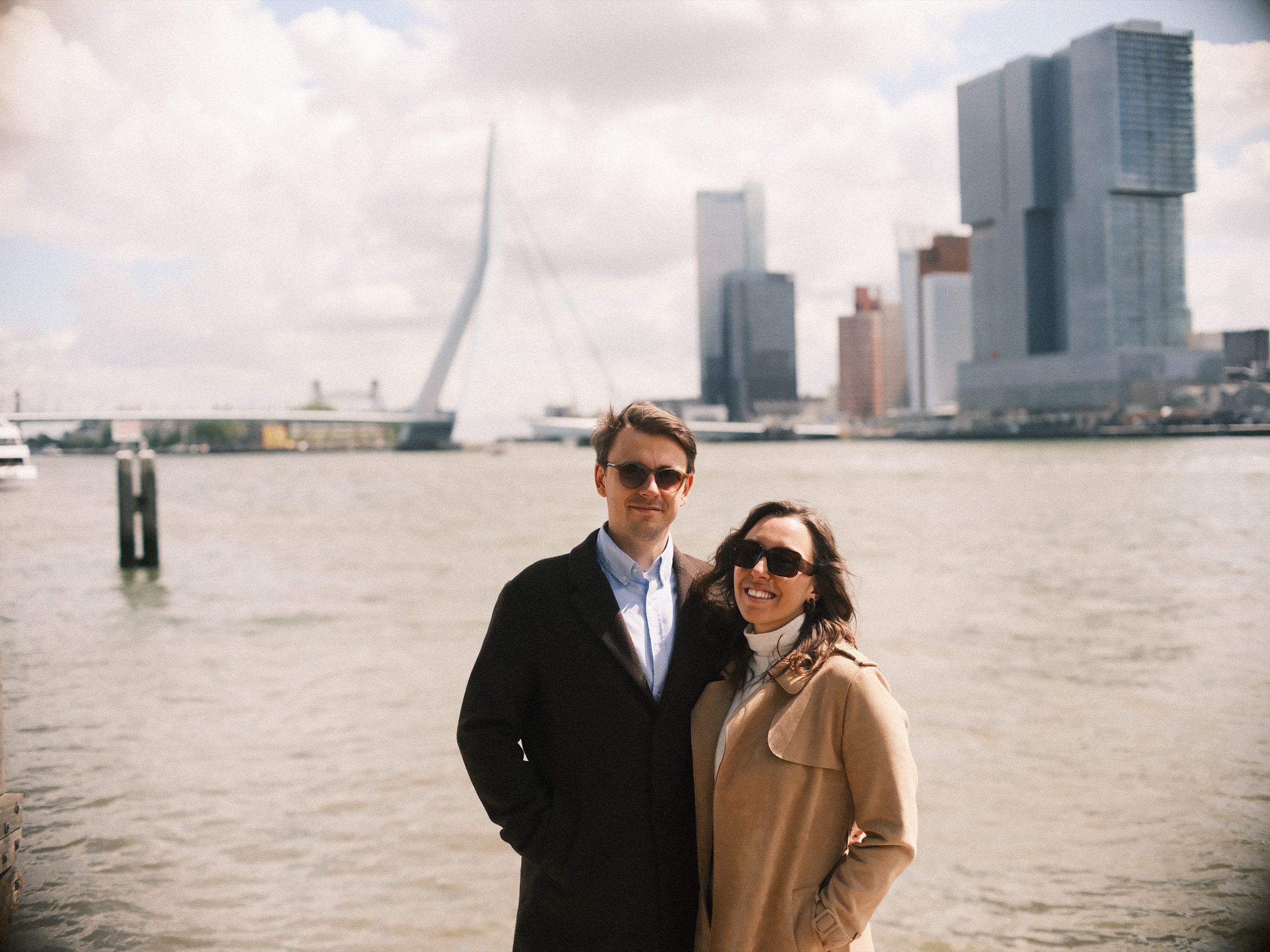 Couple in long coats and sunglasses on the bank of the Maas, the Erasmusbrug and the towers of De Rotterdam across the water