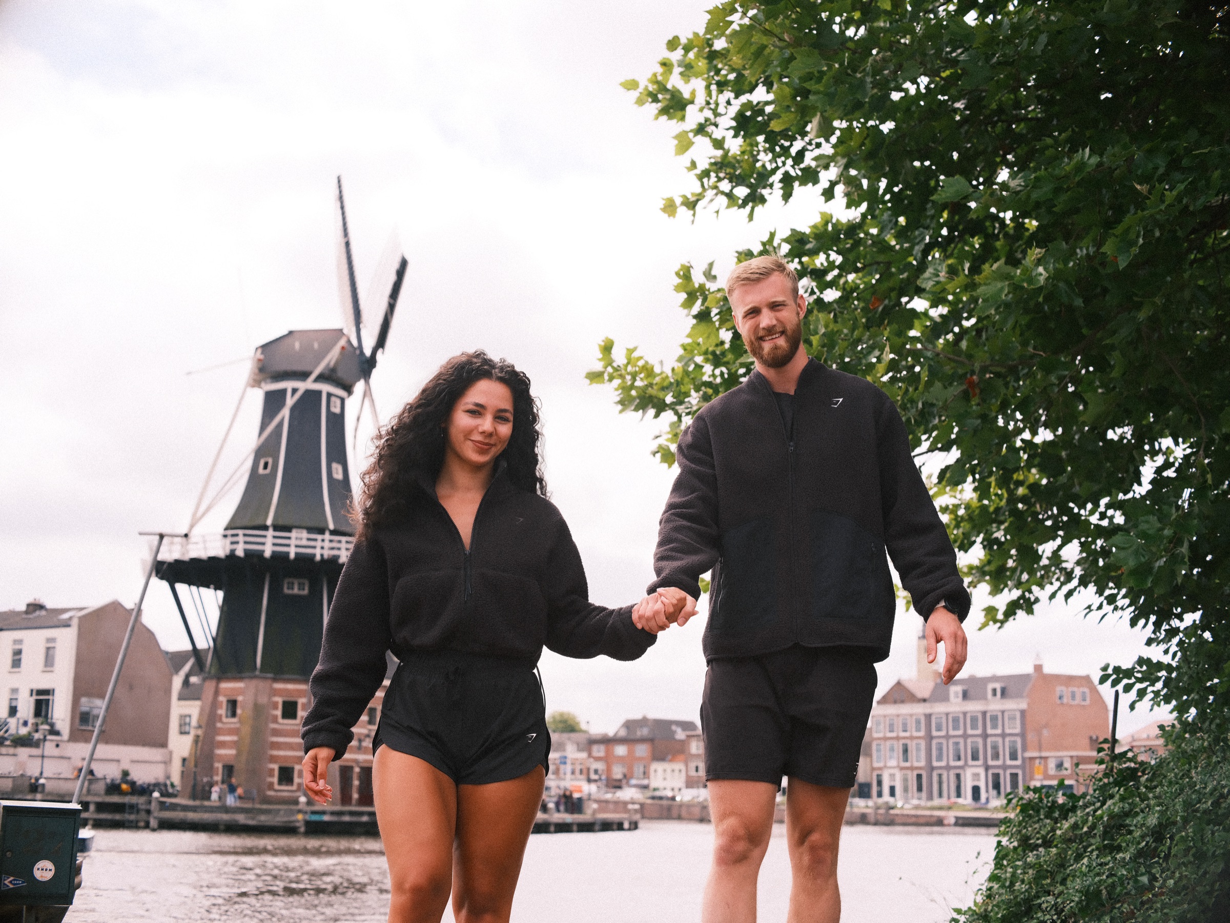 Stel in zwarte fleecevesten en korte broeken loopt hand in hand op de camera af, met molen De Adriaan aan de overkant van het Spaarne