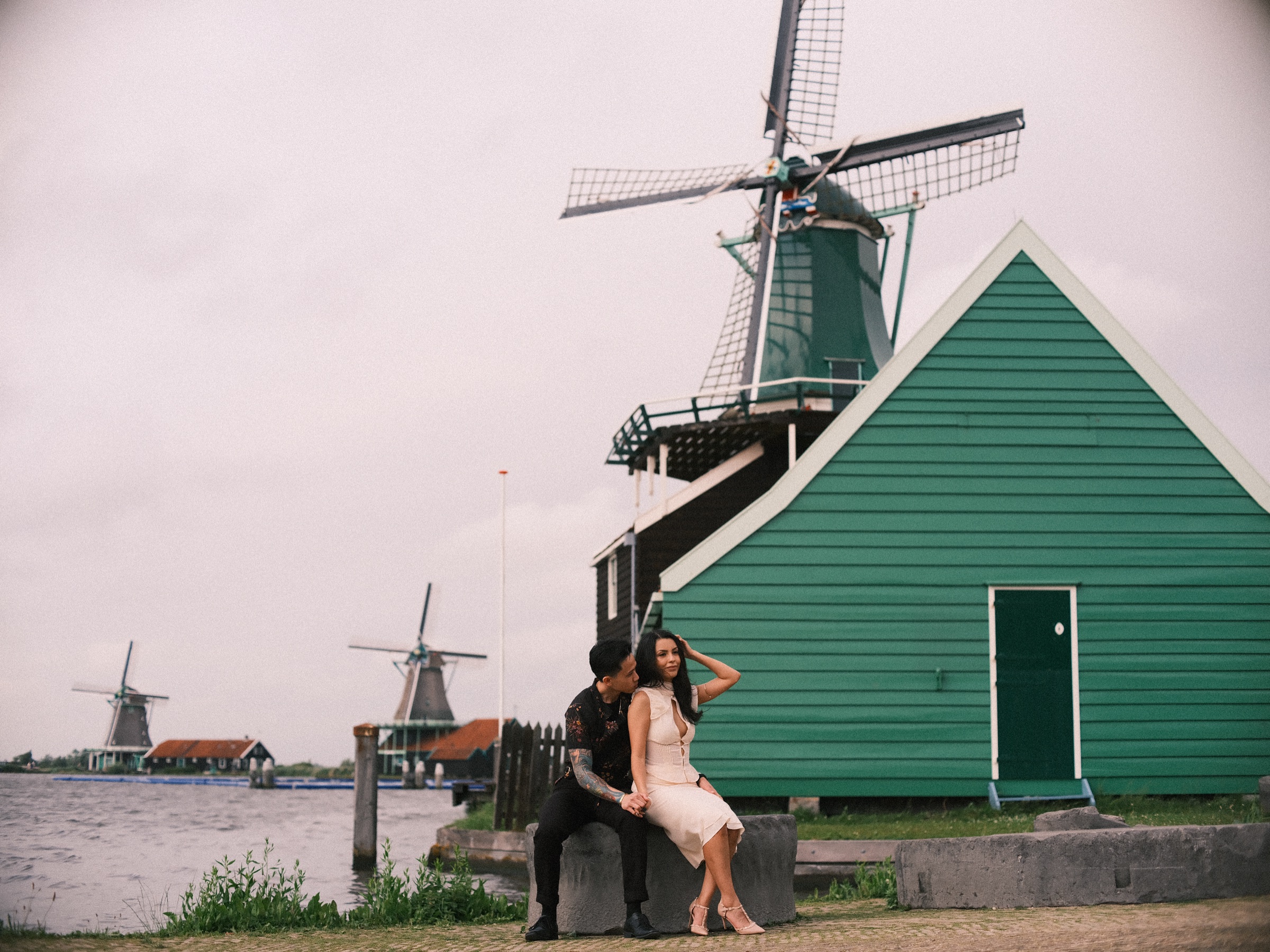 Wide view of a couple sitting on an old millstone beside a green wooden house and its windmill on the bank of the Zaan