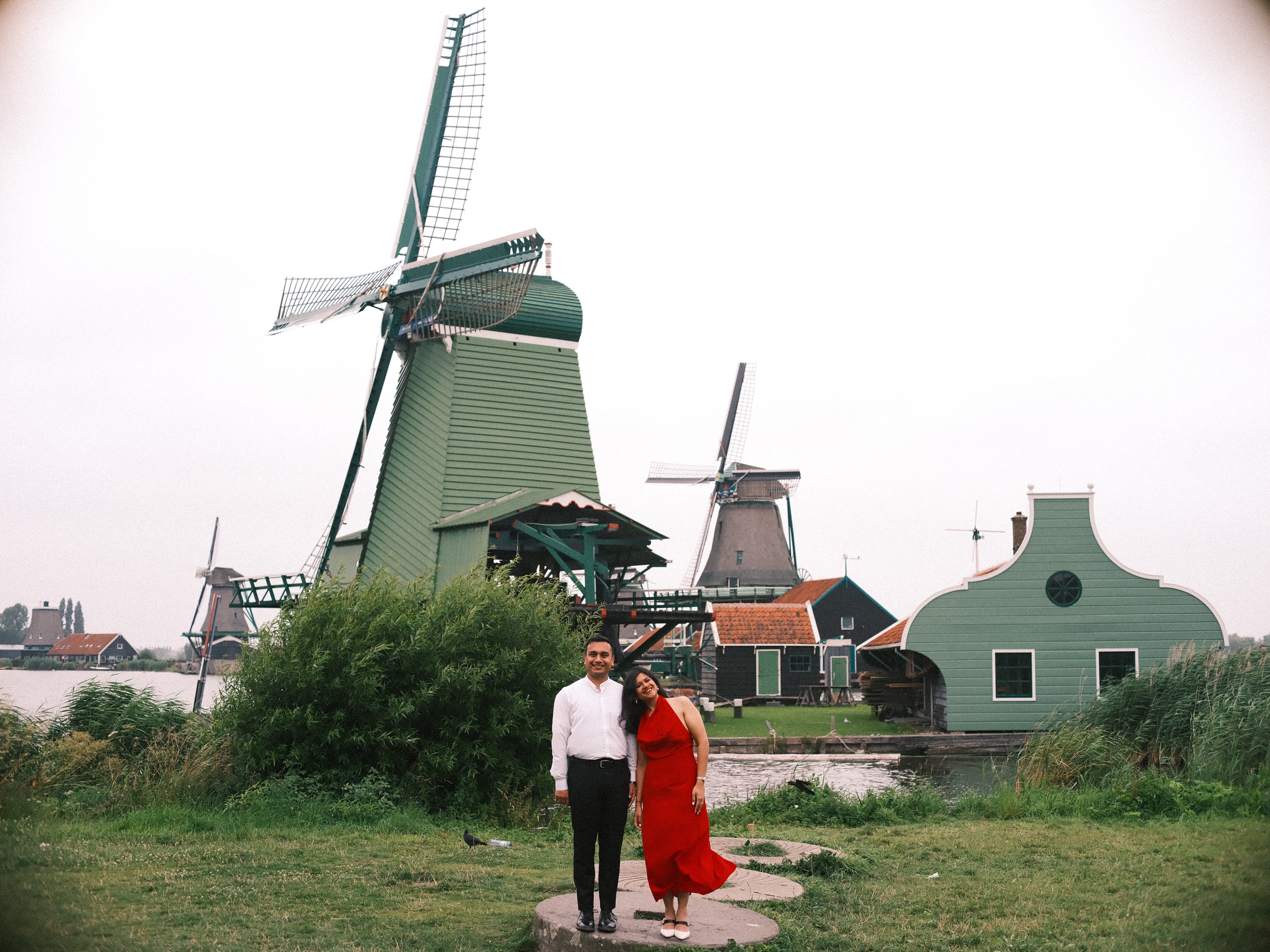Couple standing on an old millstone in the grass below a green mill at the Zaanse Schans, a light green house with a curved gable beside it