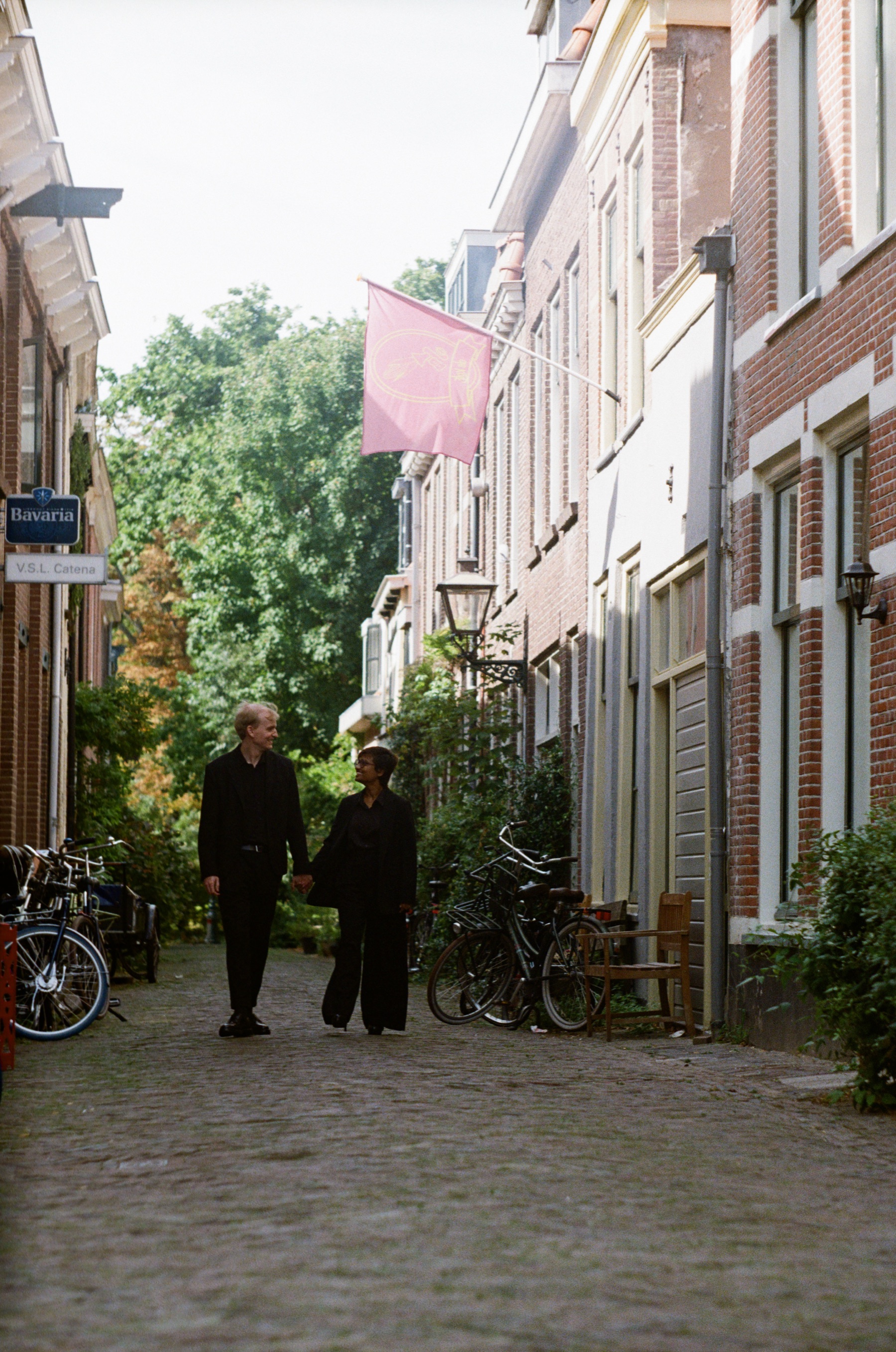 Couple in black walking down a narrow street, bikes along the walls and a pink flag overhead, color film photos from a September walk in Leiden