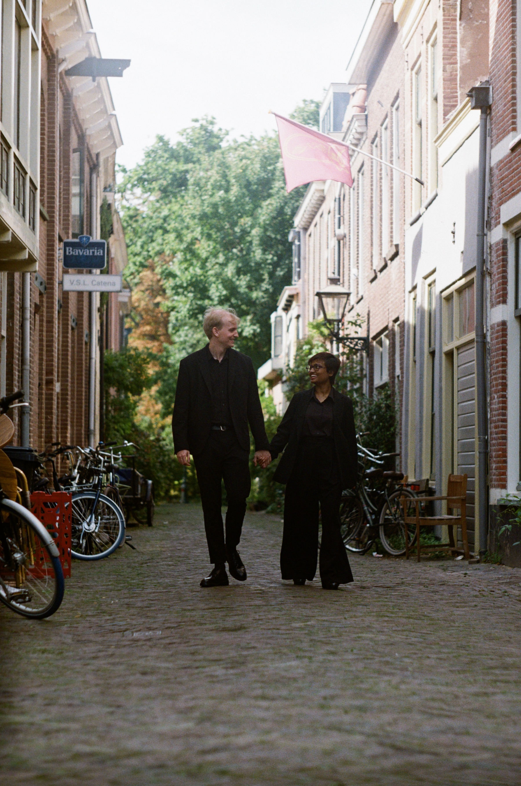 Couple in black walking toward the camera down a narrow street in Leiden, bicycles along the walls and a pink flag overhead