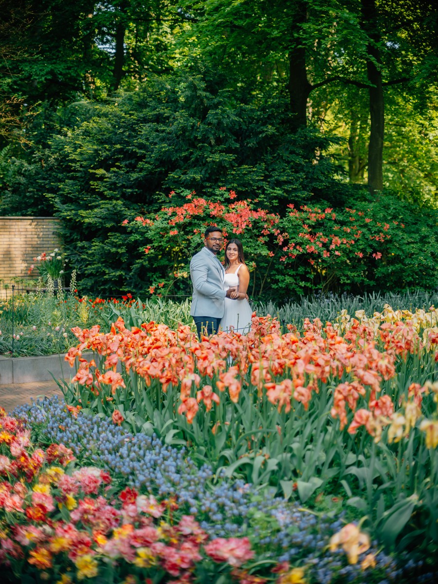 Stel staat klein achter een perk met oranje tulpen en blauwe druifjes, met een rode rododendron en bos erachter