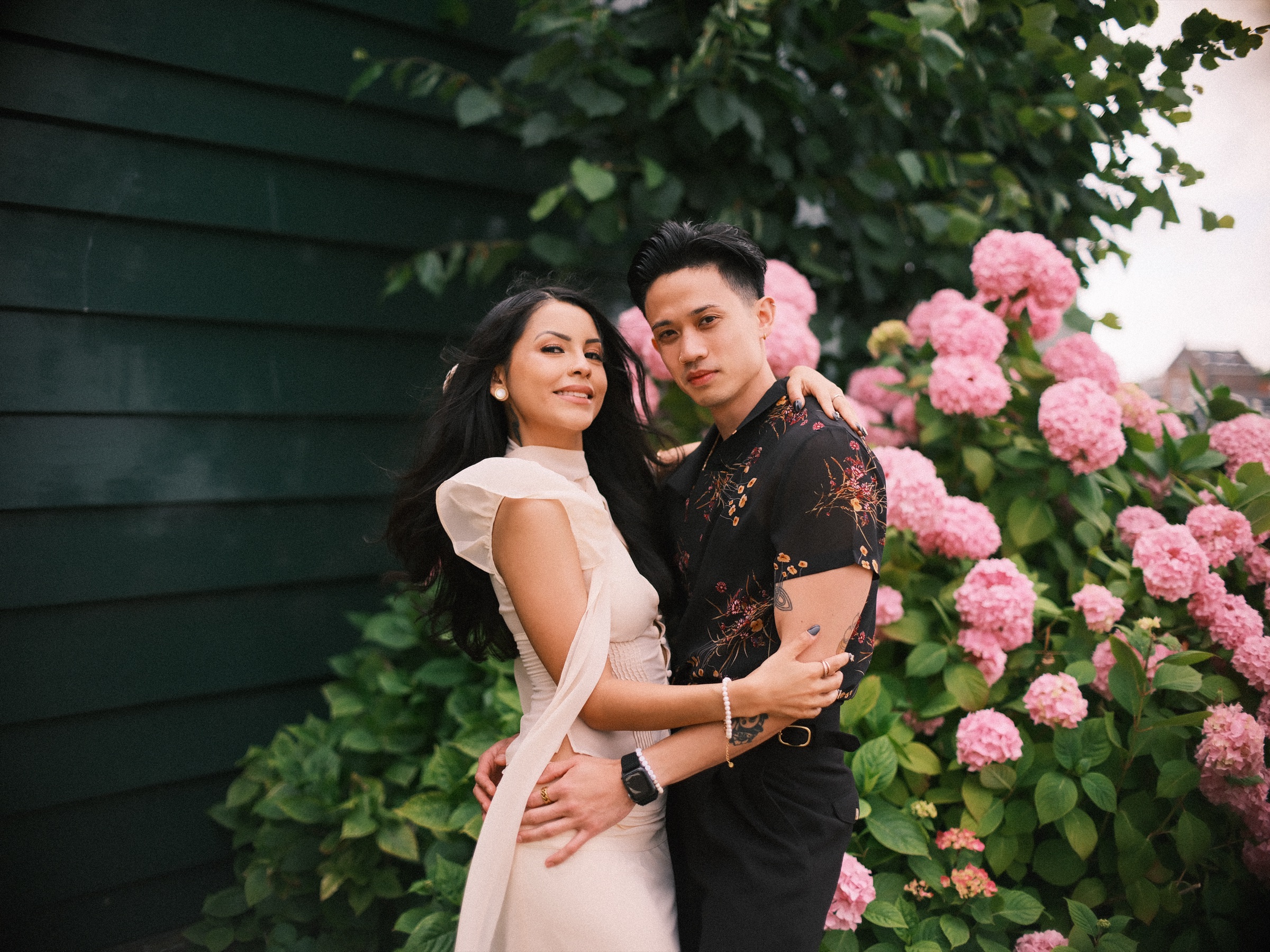 Woman in an ivory top with a long ribbon and man in a dark floral shirt hugging in front of pink hydrangeas on a dark green wooden wall