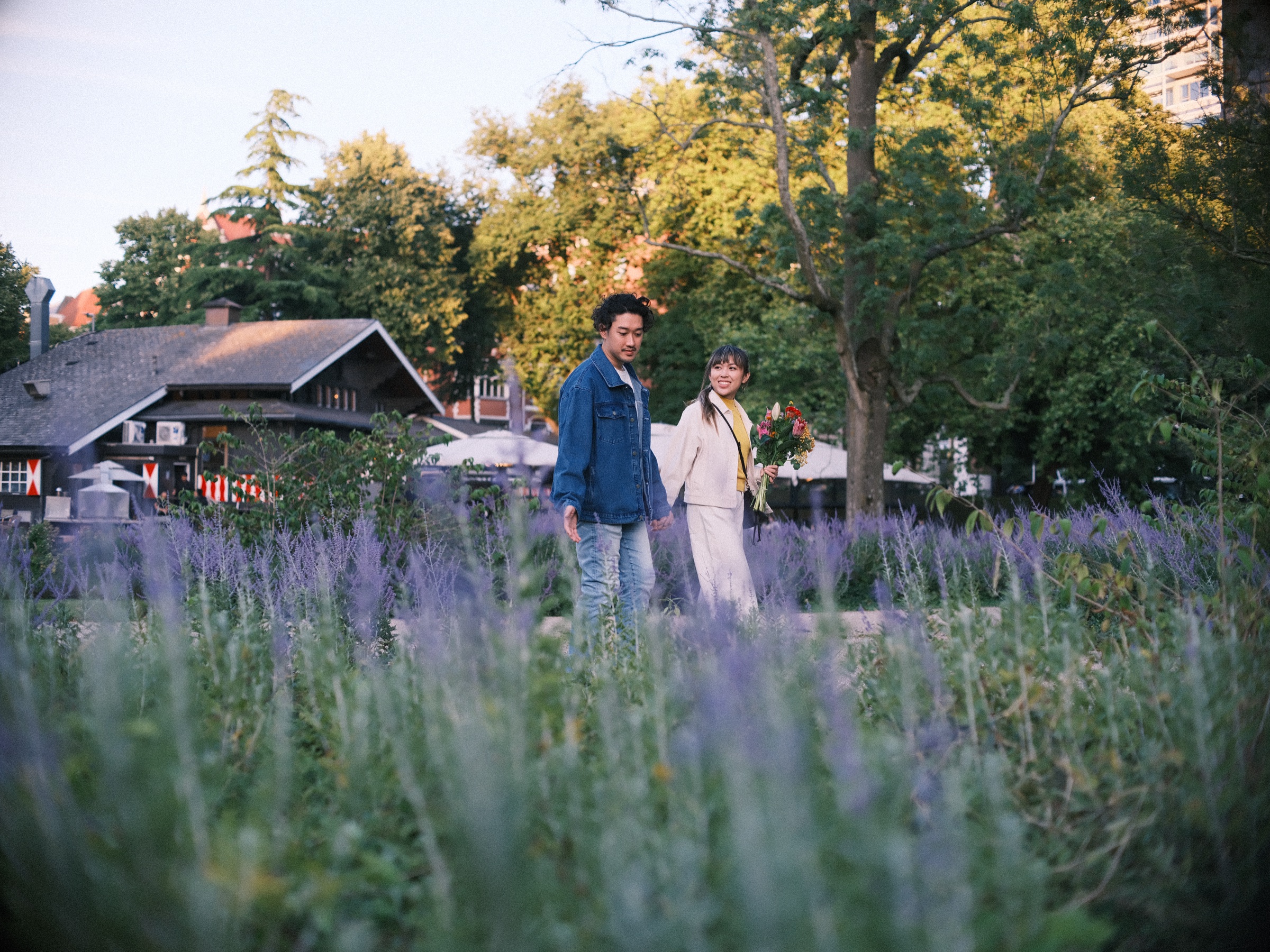 Stel tussen hoge paarse bloemen in Het Park, met donkere houten gebouwen en de parasols van een terras op de achtergrond