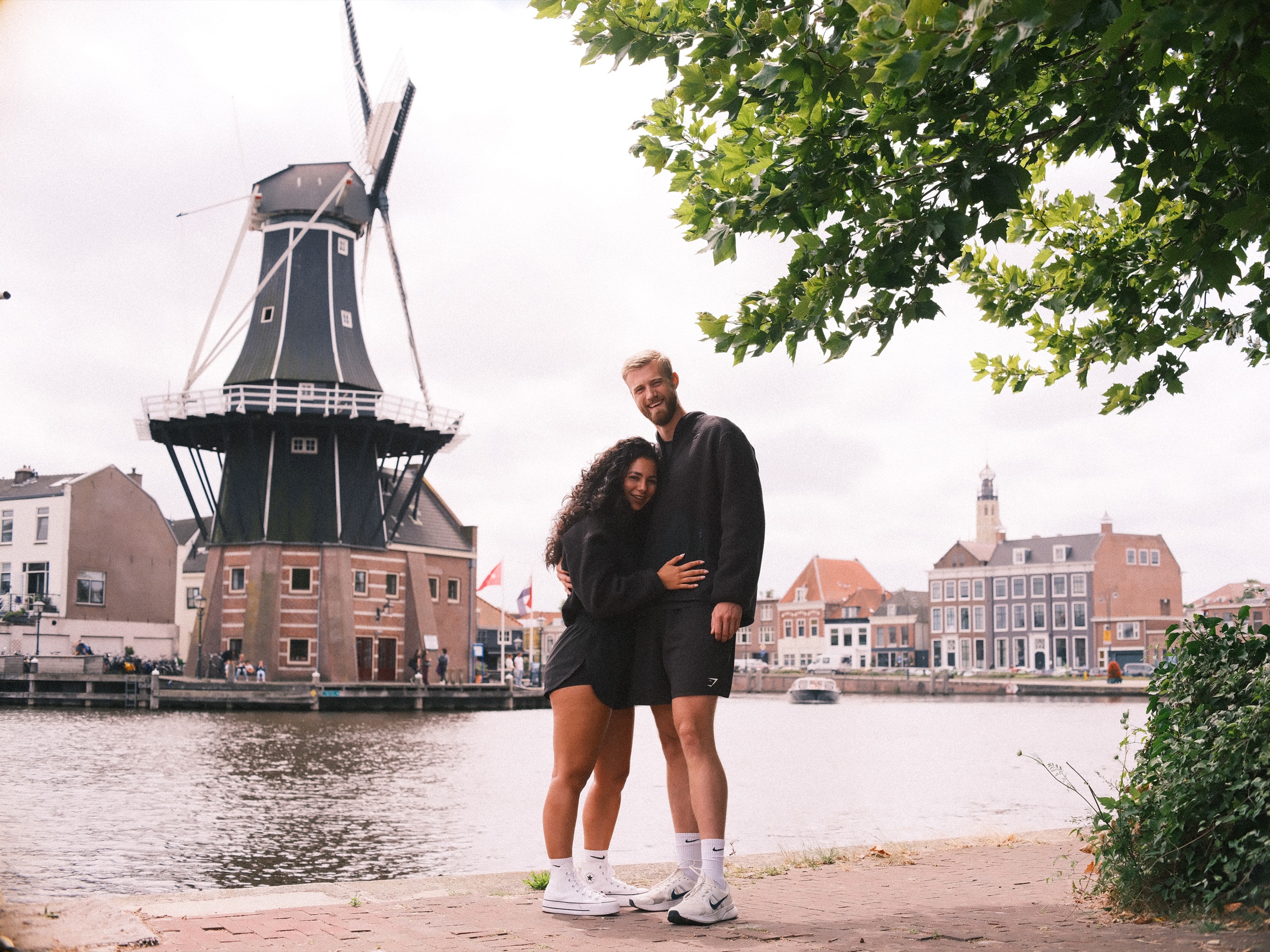 Stel in bijpassend zwart dicht tegen elkaar op de kade tegenover molen De Adriaan, met een lommerrijke boom boven het water