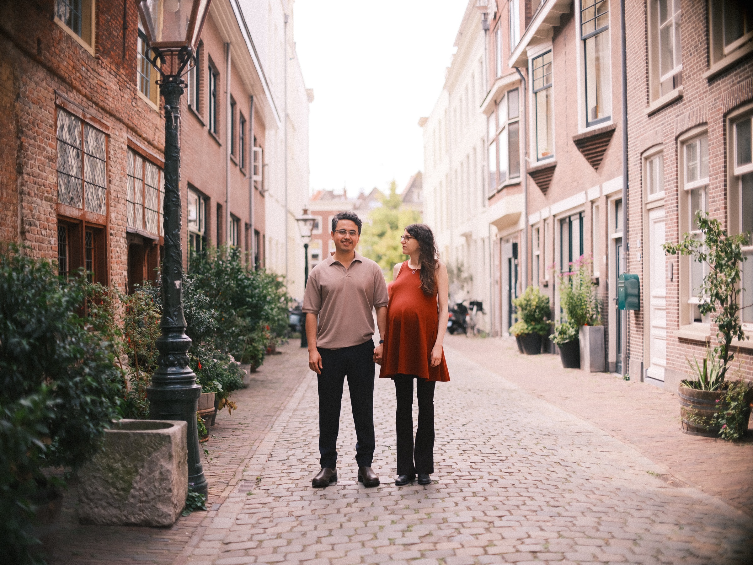 Couple standing in a long cobbled lane with an old lamppost and potted plants, brick houses on both sides, on a summer photo walk in Leiden