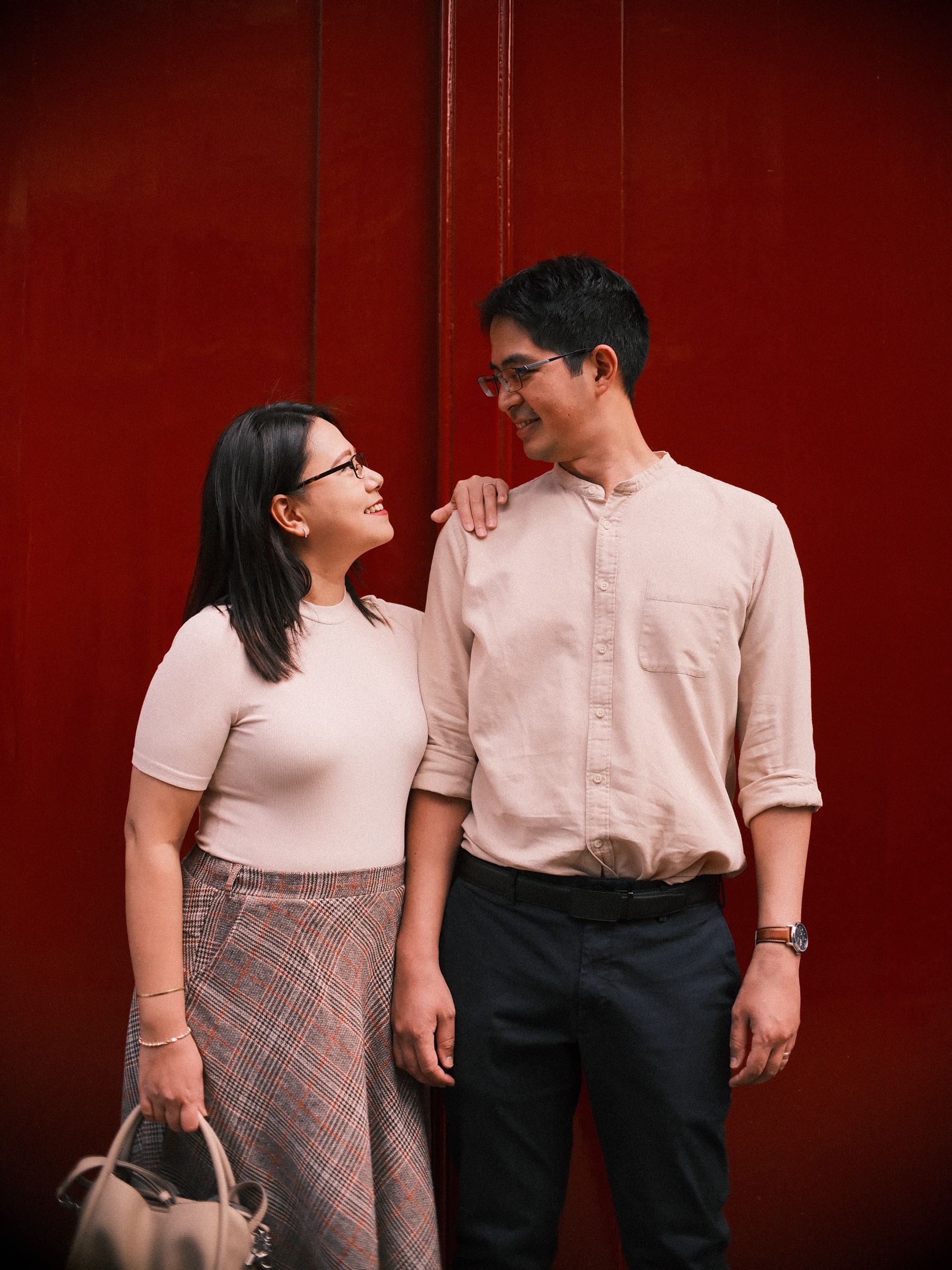 Couple smiling at each other in front of a tall red door in Leiden, her hand on his shoulder, both dressed in soft cream and pink