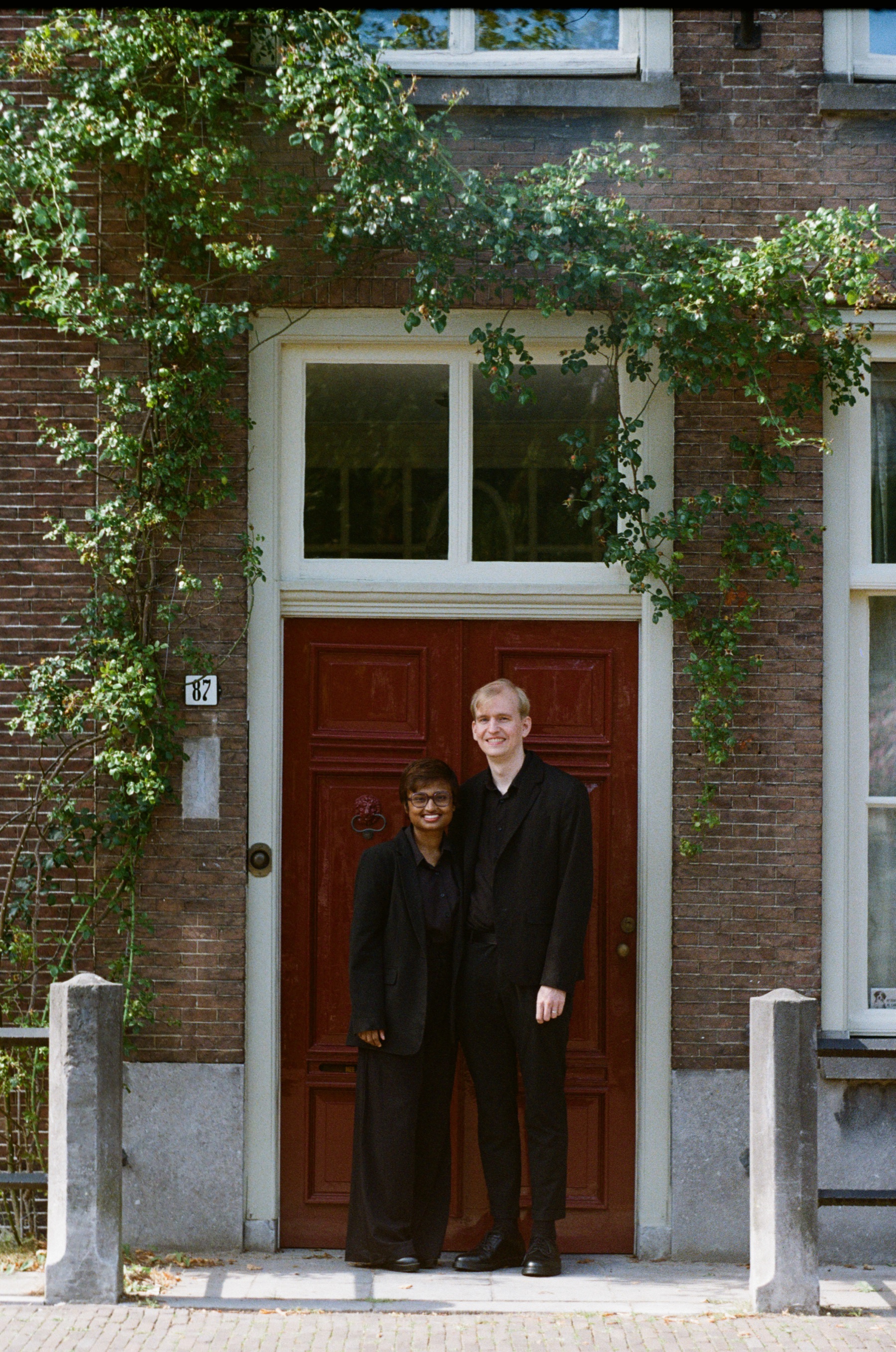 Couple in black side by side at a red front door framed by ivy and white window frames, two stone posts on the brick pavement