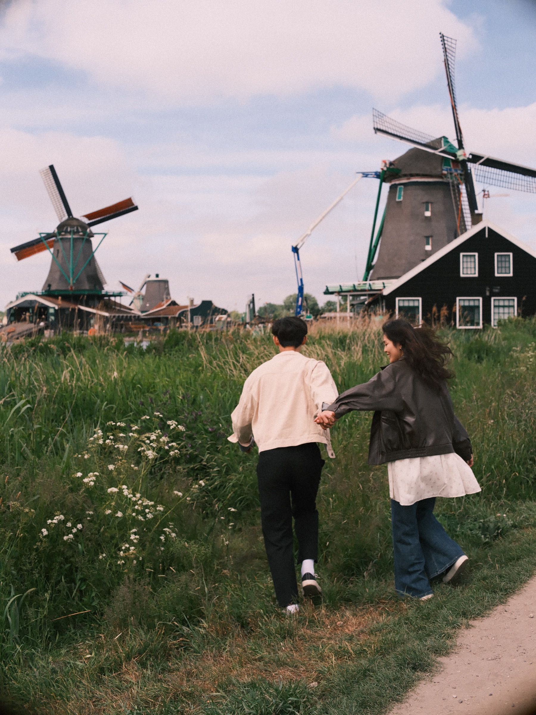 Couple running hand in hand through long grass and white wildflowers toward two windmills, seen from behind on a breezy day