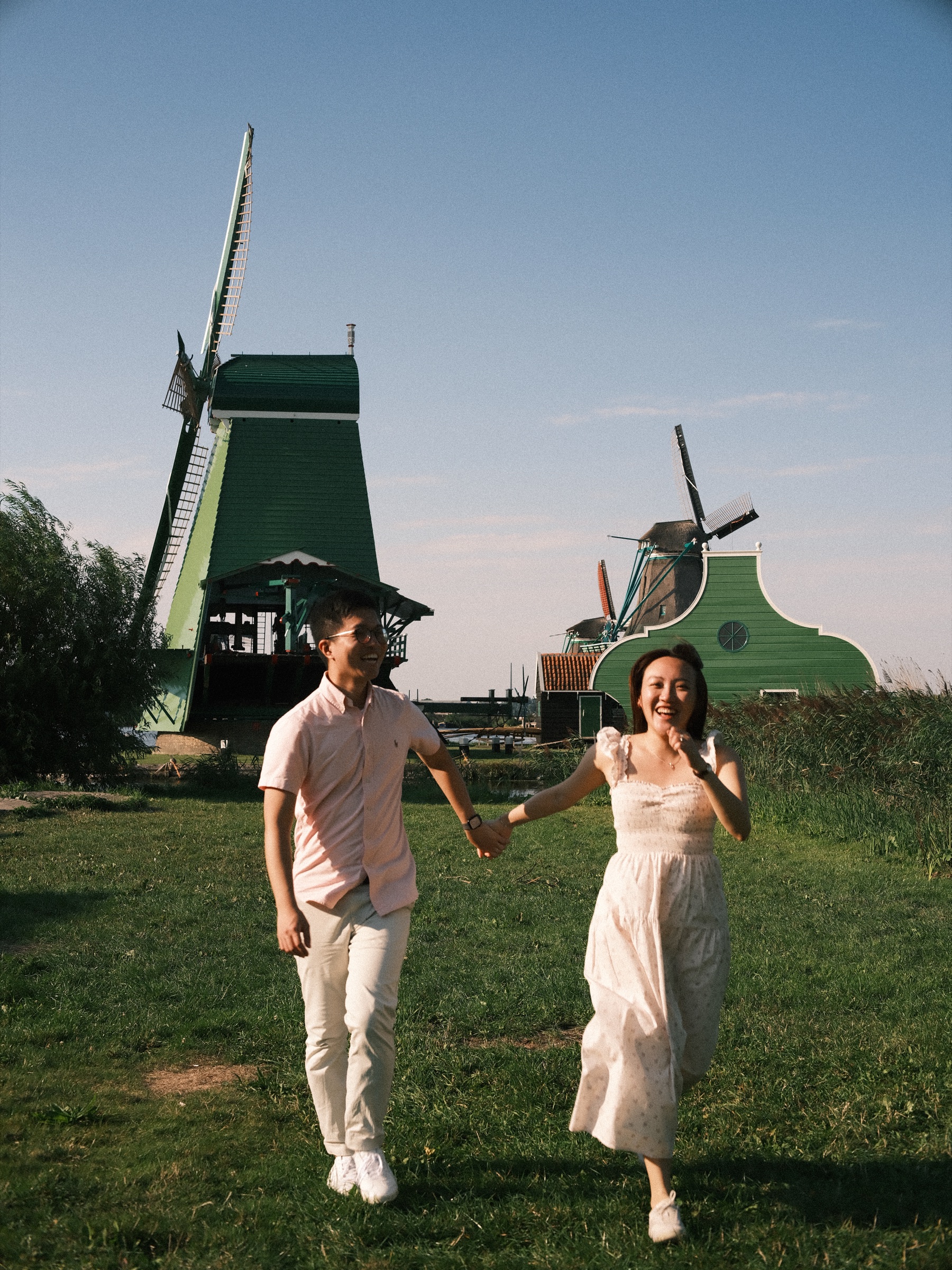Couple running hand in hand across the grass and laughing, the windmills of the Zaanse Schans against a blue sky