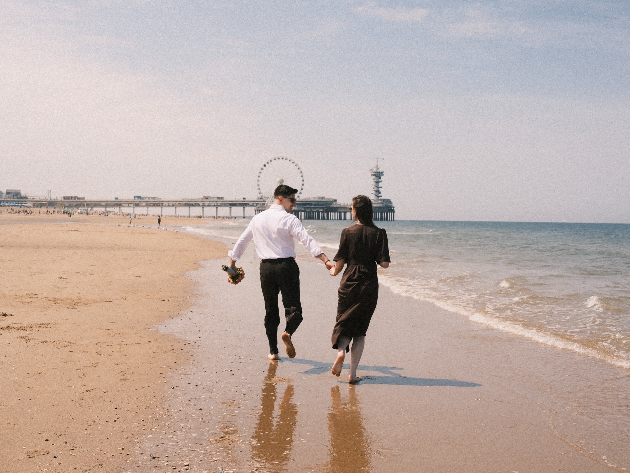 Engaged couple running hand in hand along the tide line toward the Pier and its Ferris wheel on Scheveningen beach