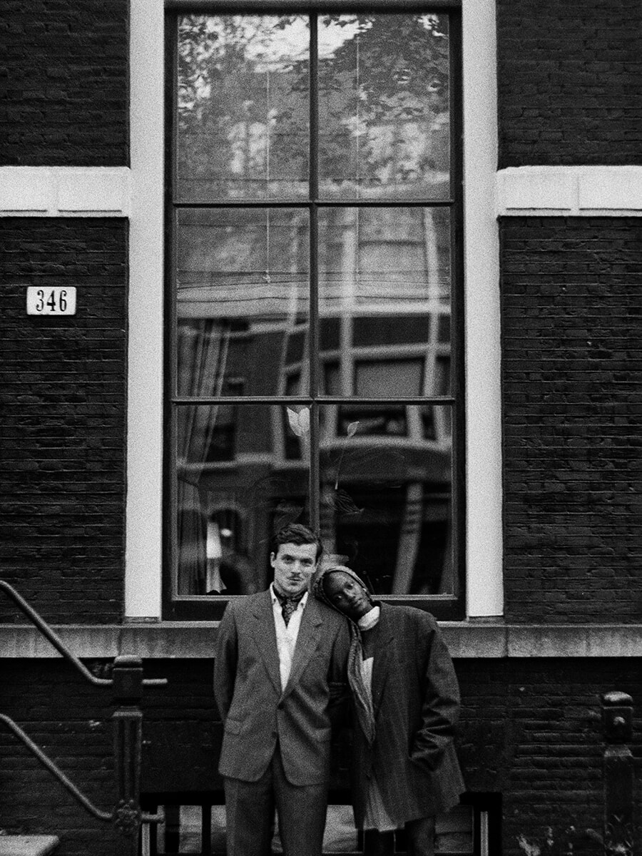 Resting her head on her partner’s shoulder, a woman stands beneath a tall window reflecting the buildings opposite