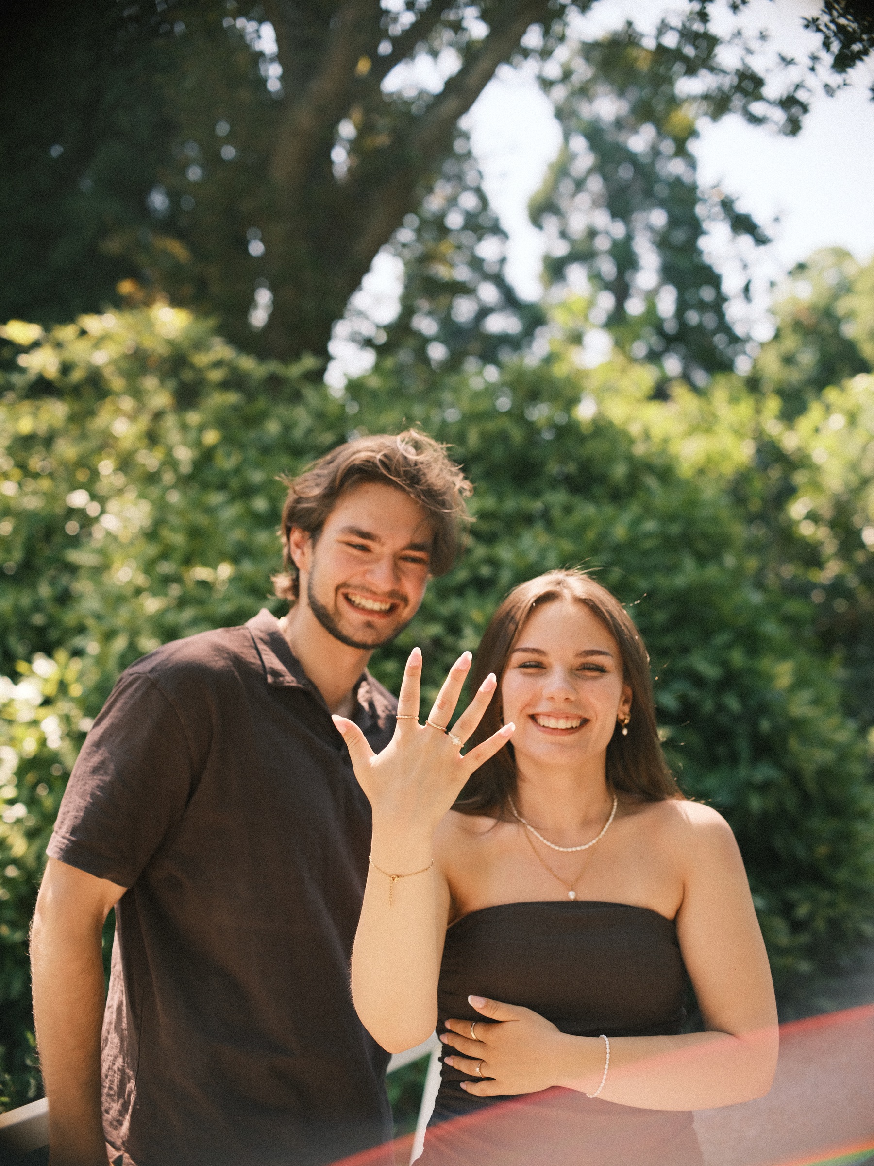 Smiling together, she holds up her hand to show the new ring, his arm around her waist, after the surprise proposal at Trompenburg