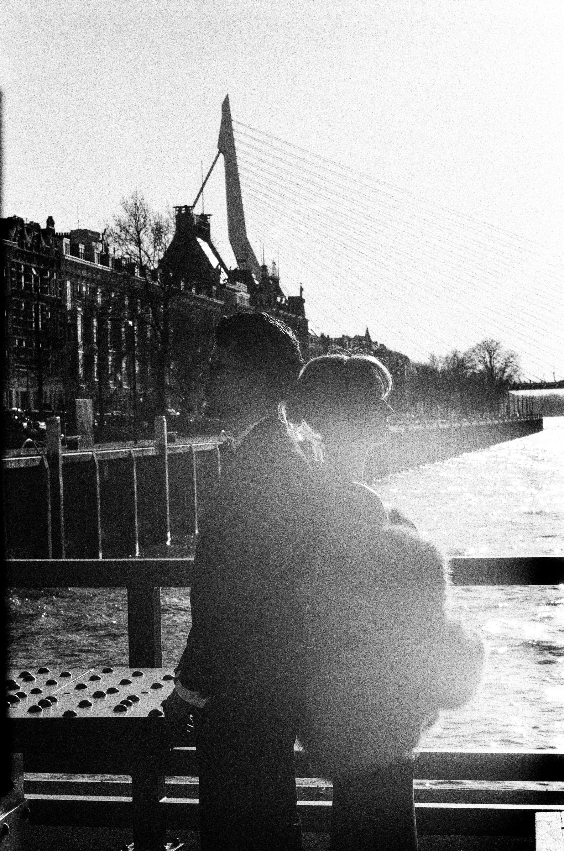 Couple in silhouette against the sun on a quay by the Maas, the Erasmusbrug and old houses behind them, a soft flare over her fur coat