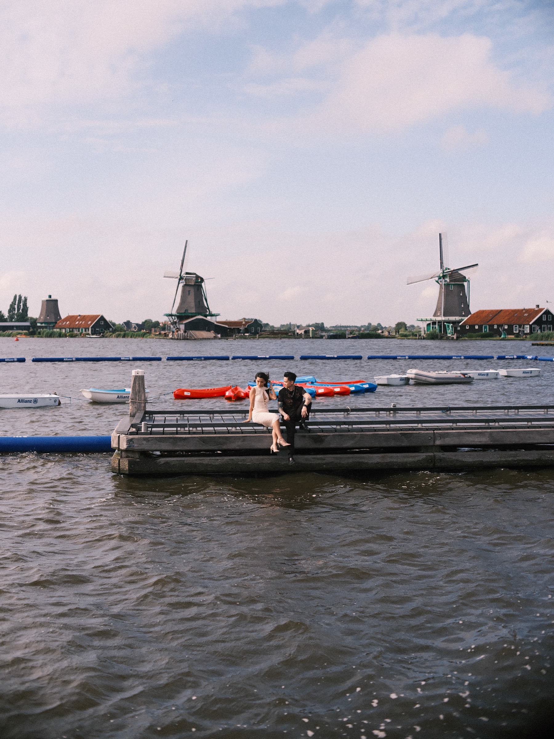 Couple sitting on the edge of a wooden jetty in the Zaan, red and blue boats moored behind them and two windmills on the bank