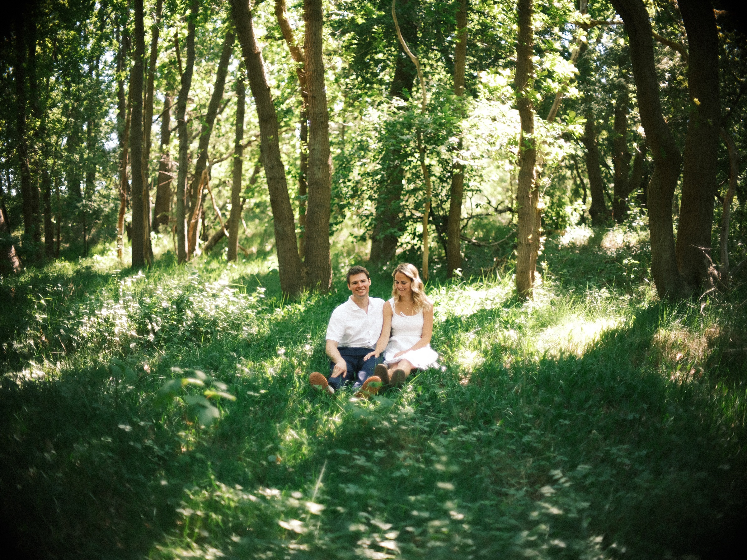 Stel zit dicht bij elkaar in hoog groen gras tussen slanke bomen, klein in een wijd beeld van het bos in de duinen