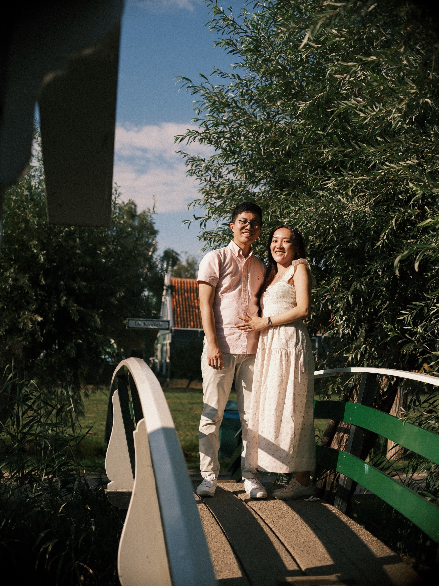 Woman resting a hand on her fiancé's chest on a small bridge with white railings, green leaves around them in the late sun