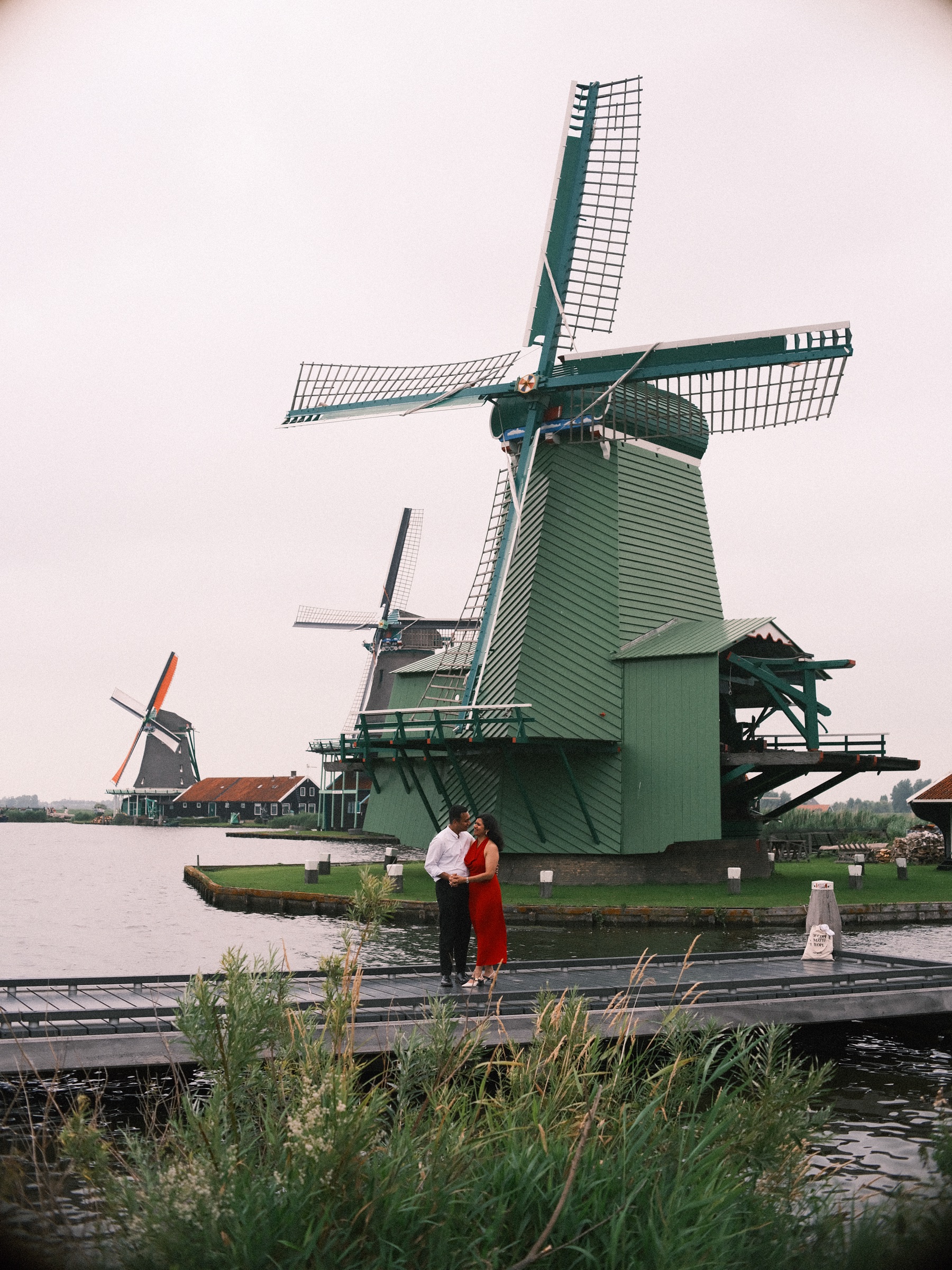 Couple standing small on a wooden jetty below the big green windmill, tall reeds in front and the water of the Zaan around them