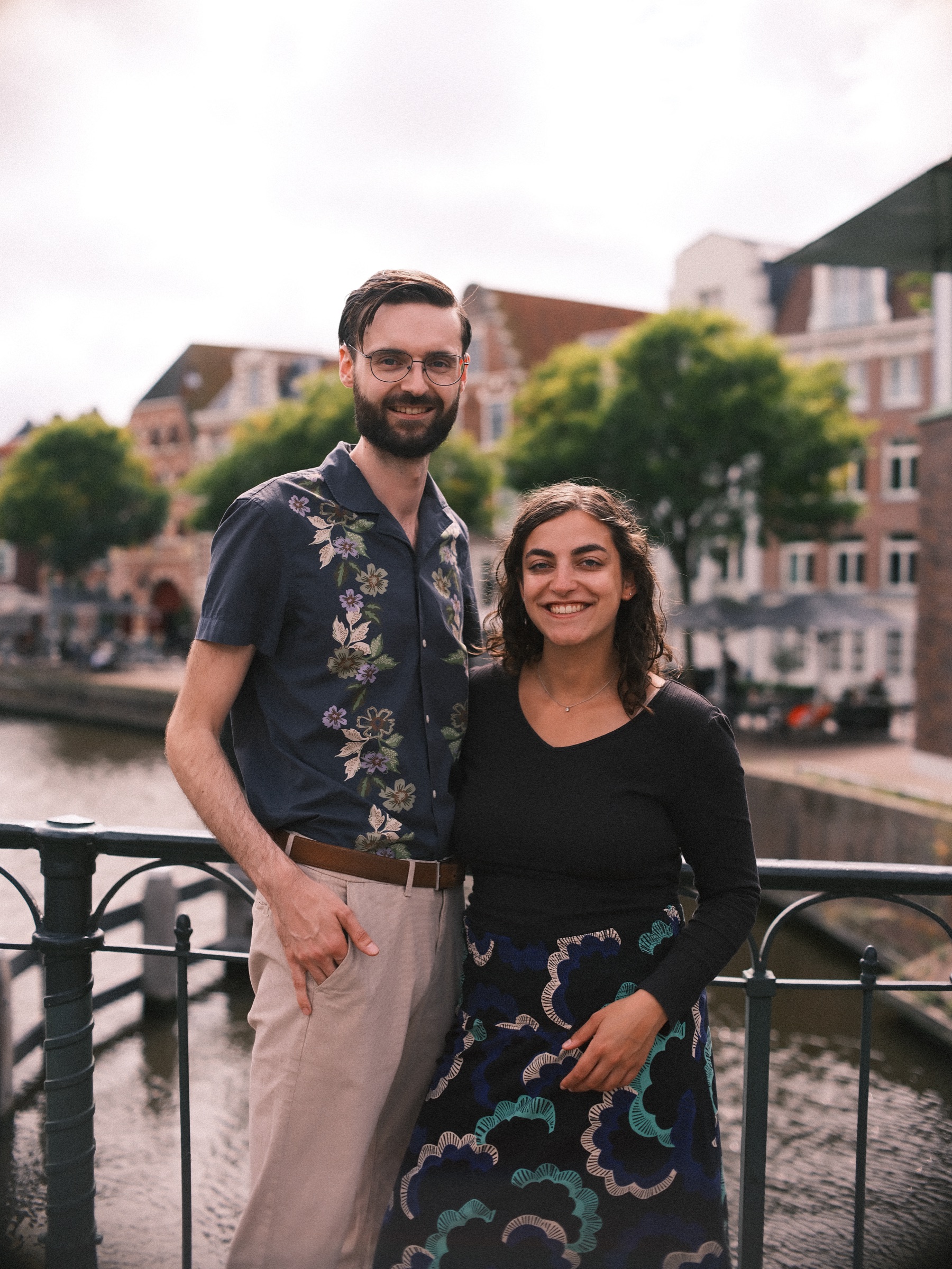 Stel lacht naar de camera aan het water, hij in een blauw overhemd met bloemen en zij in een zwart topje en een donkerblauwe rok