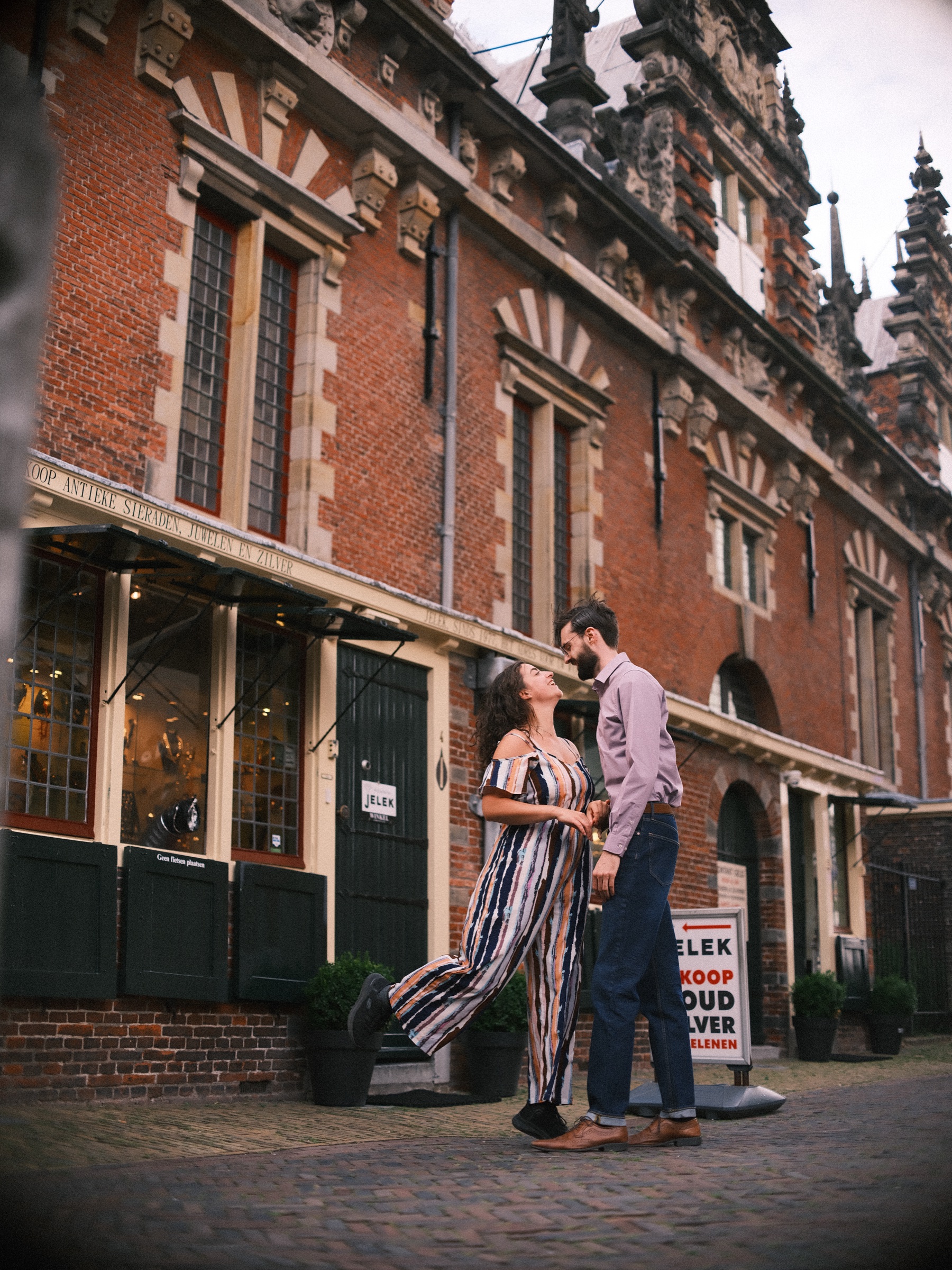 Vrouw in een gestreepte jumpsuit tilt een voet op en lacht naar haar partner bij de Vleeshal op de Grote Markt in Haarlem, onder versierde gevels
