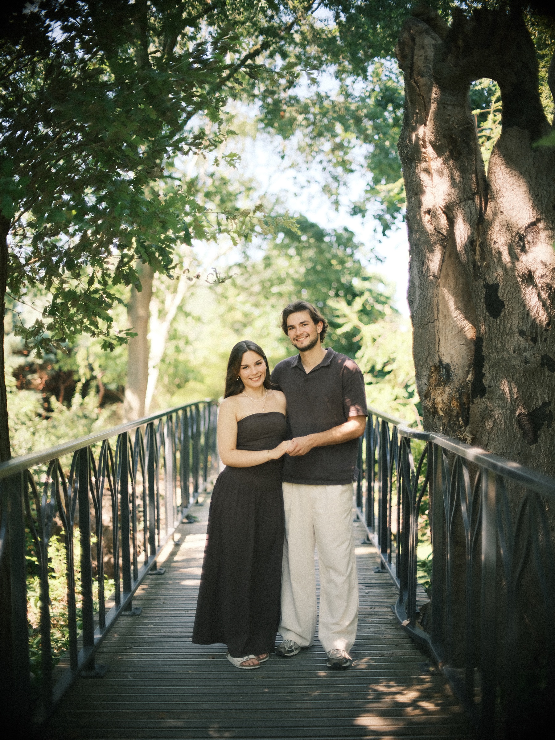 Couple standing on a dark iron bridge beside a huge hollow tree in the Trompenburg gardens, a green photo spot in Rotterdam