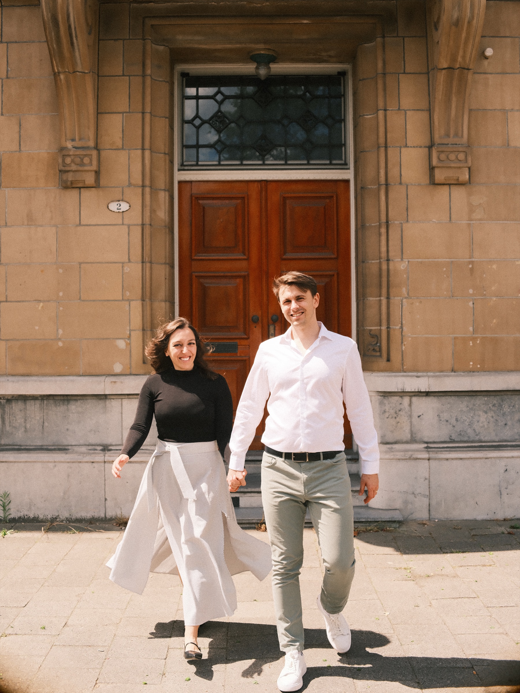 Stepping out hand in hand from a tall wooden doorway in the Scheepvaartkwartier, she in a pale pleated skirt during their engagement shoot