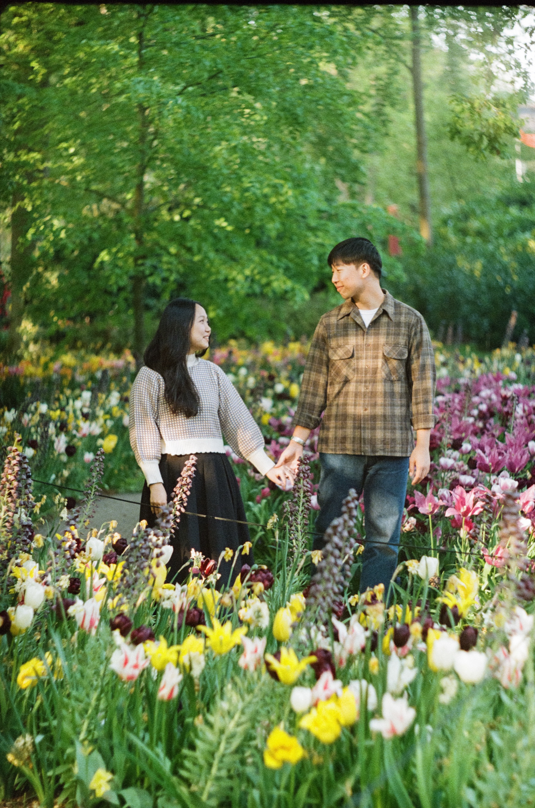 Man in a plaid shirt and woman in a long black skirt strolling hand in hand past purple and yellow flowers