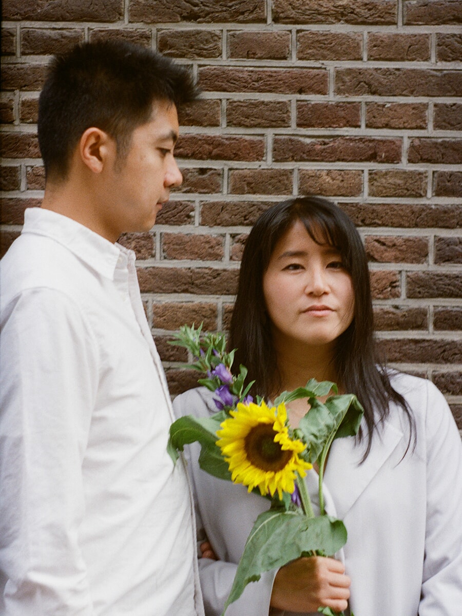 Holding a sunflower and purple flowers, a woman stands beside her partner against a brick wall in a colour film portrait